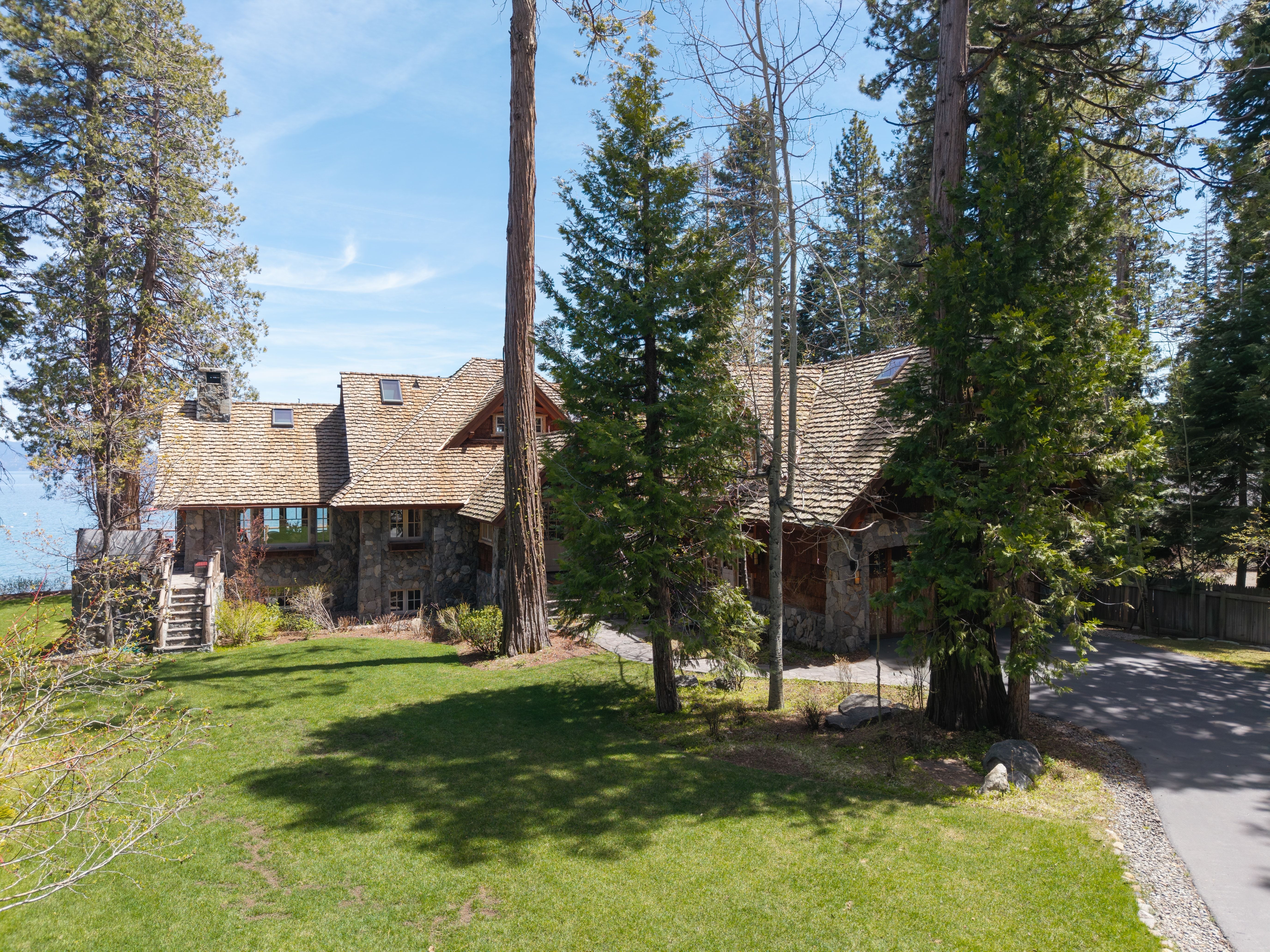 This charming, multi-story home features classic mountain architecture with a combination of rough-hewn stone siding and cedar shake roofing. Nestled amongst lush, mature evergreens, the property includes a well-manicured lawn and a private paved driveway, creating an inviting and secluded atmosphere. A stone staircase leads toward the main entry area, suggesting a refined, rustic aesthetic that complements the surrounding natural landscape.