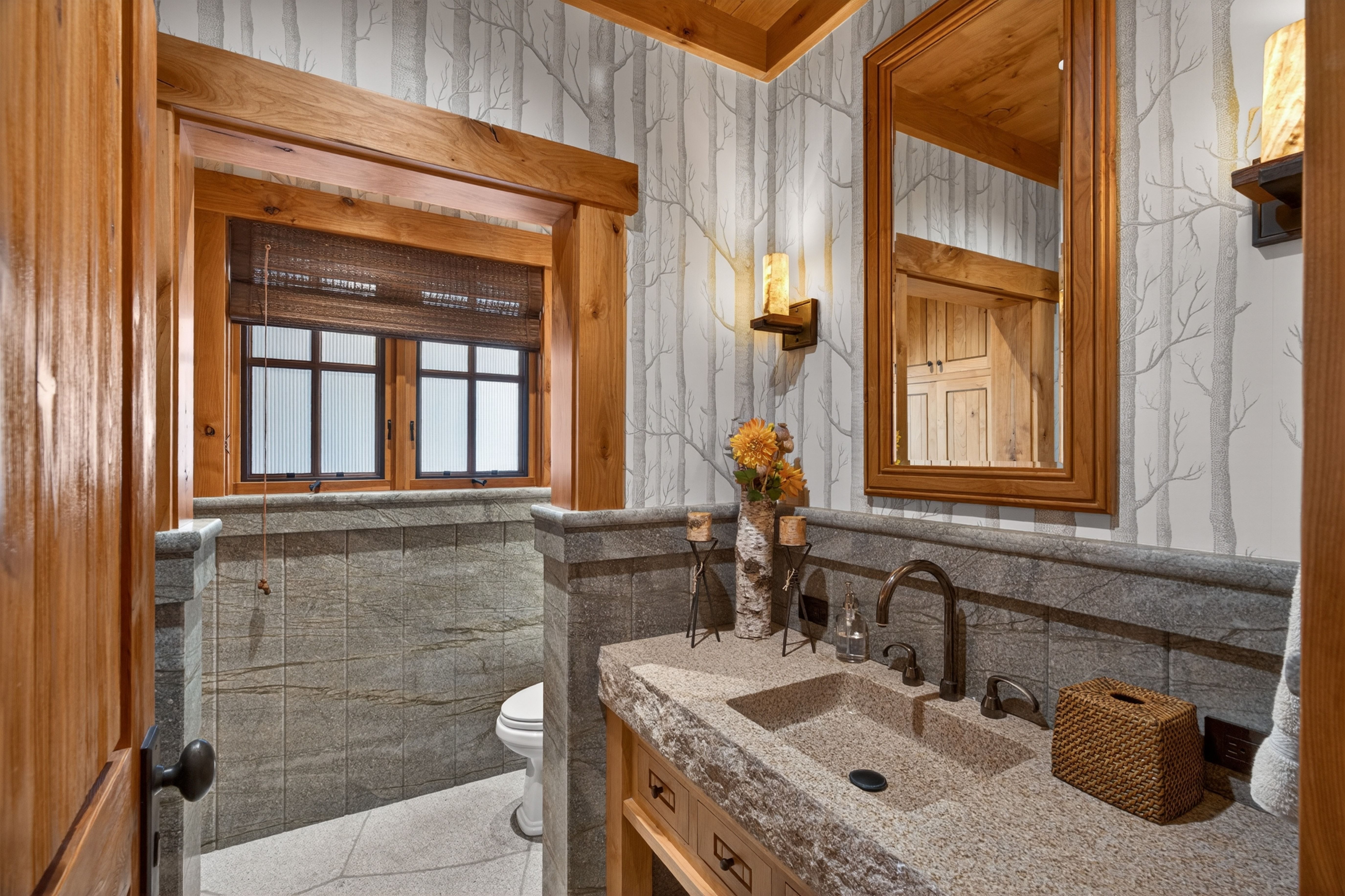 This rustic powder room features a unique stone-carved sink integrated into a granite countertop and vanity. The striking woodland-themed wallpaper, combined with warm natural wood trim, fixtures, and candle-style wall sconces, creates a cozy, retreat-like atmosphere. The space is well-defined by stone wall paneling and finished with precise craftsmanship, ideal for a mountain home setting.
