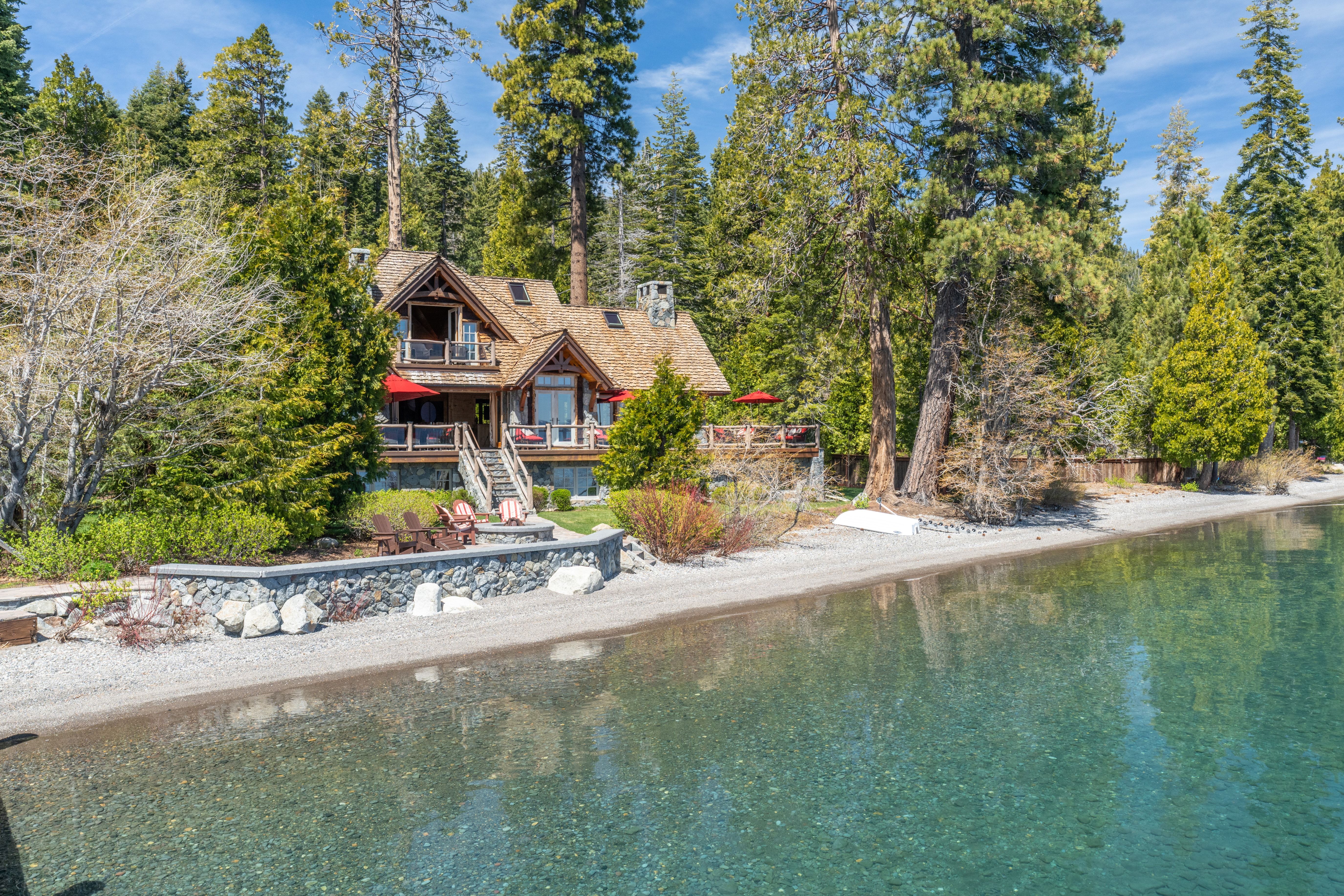 This scenic lakefront property features a multi-level wooden cabin-style home nestled among tall pine trees. The house includes a spacious deck with red umbrellas, stone accents, and a private stone-walled patio area right along the clear, pebbled shoreline. The tranquil water and lush forest setting highlight the home's premier position for lakeside relaxation.