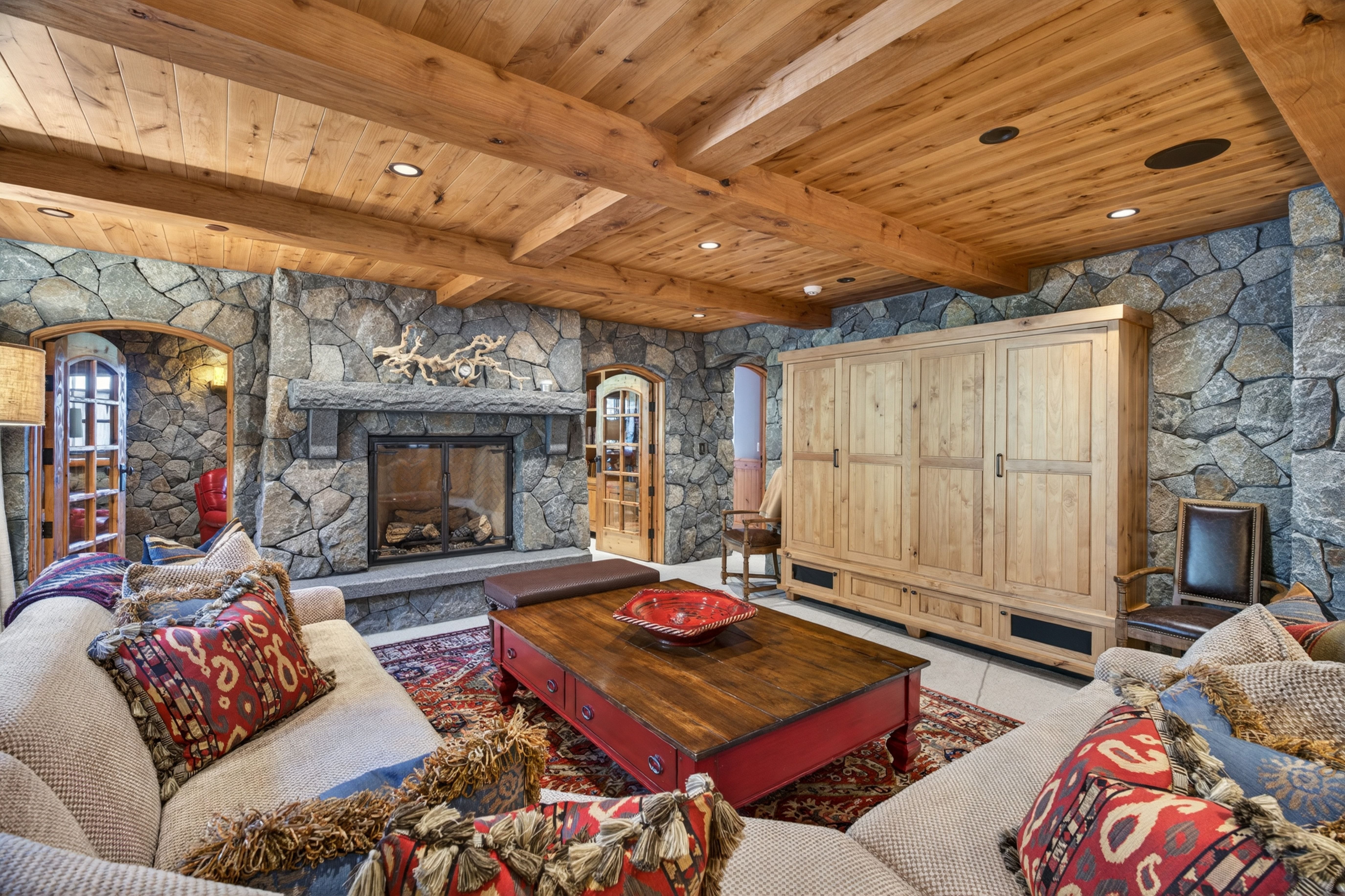 This cozy living room features a rustic cabin aesthetic with a prominent stone fireplace, exposed wood beamed ceilings, and natural stone accent walls. The space is furnished with overstuffed couches, a large wooden coffee table with red accents, and a traditional patterned rug. A large, light-colored wooden cabinet provides substantial storage, completing the warm and inviting mountain lodge atmosphere.