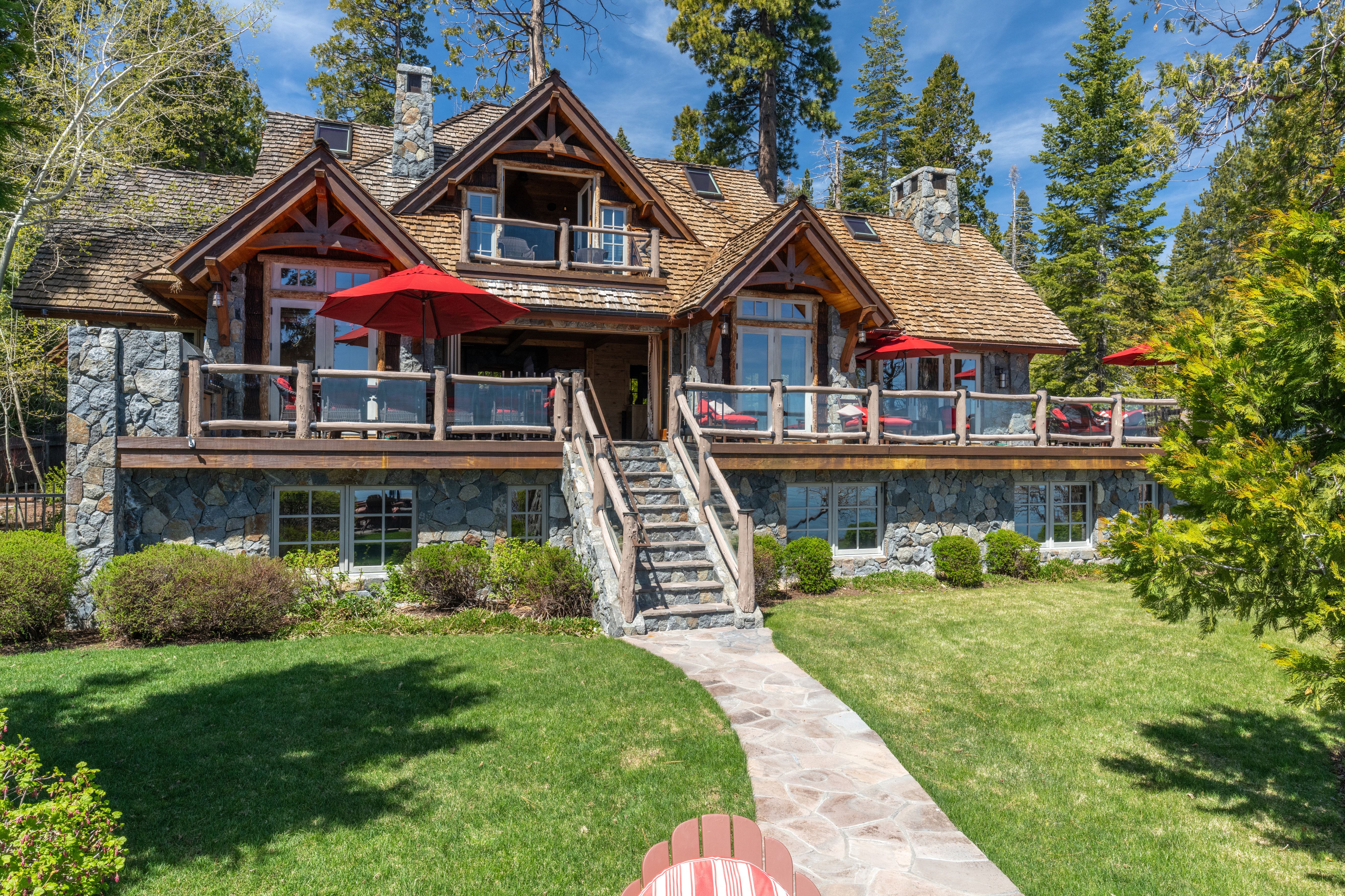 This exquisite mountain estate features a stunning timber and stone facade, highlighted by a spacious raised deck with multiple red umbrellas and ample seating. A centrally located stone staircase connects the deck to a manicured lawn and a stone walkway. The home offers a classic, upscale alpine lodge aesthetic set against a backdrop of mature pine trees.