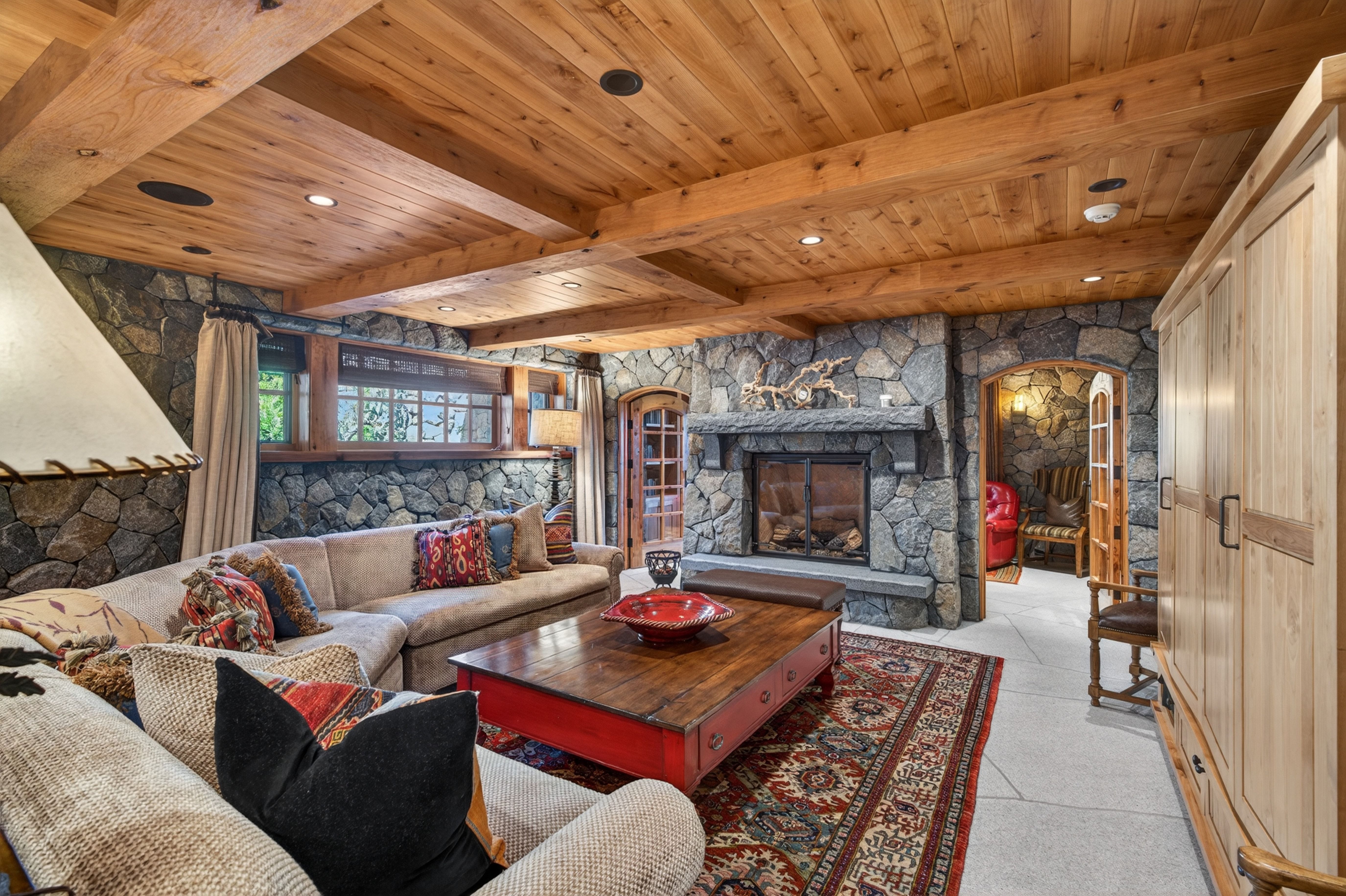 This cozy, rustic-style living room features impressive stone walls, exposed timber beams on the ceiling, and a prominent stone fireplace. A large sectional sofa, a sturdy wooden coffee table, and an intricate area rug create a warm and inviting atmosphere. The space exudes charm and character, perfect for relaxation in a mountain or lodge-inspired home.