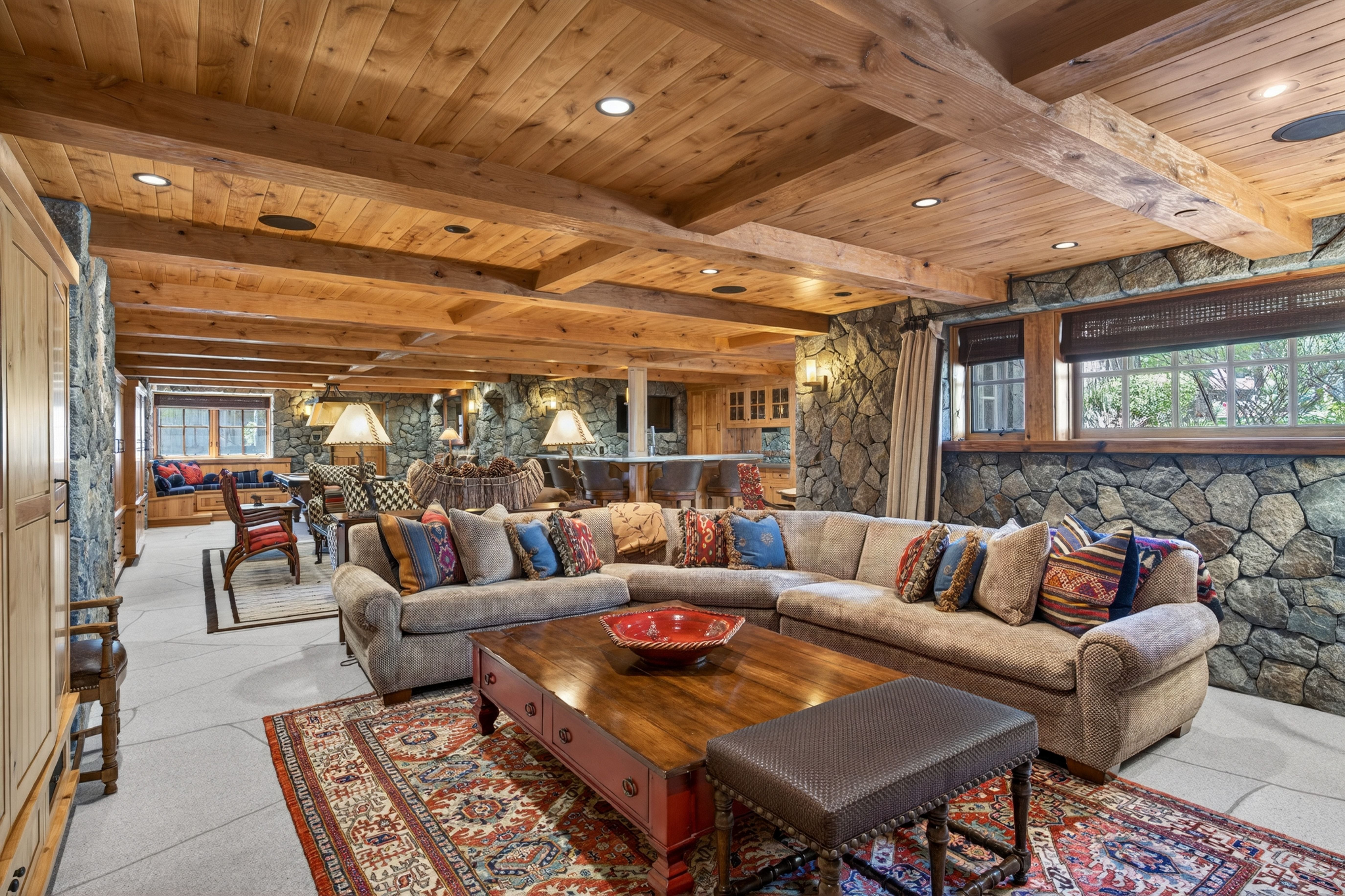 This spacious and rustic-style living room features warm wooden ceiling beams and natural stone accent walls that create a cozy atmosphere. A large, neutral-toned sectional centerpiece is complemented by an ornate area rug, a polished wooden coffee table, and an ottoman, making it ideal for relaxing or entertaining. The open layout extends to a bar area in the back, while recessed lighting emphasizes the craftsmanship of the timber-framed structure.