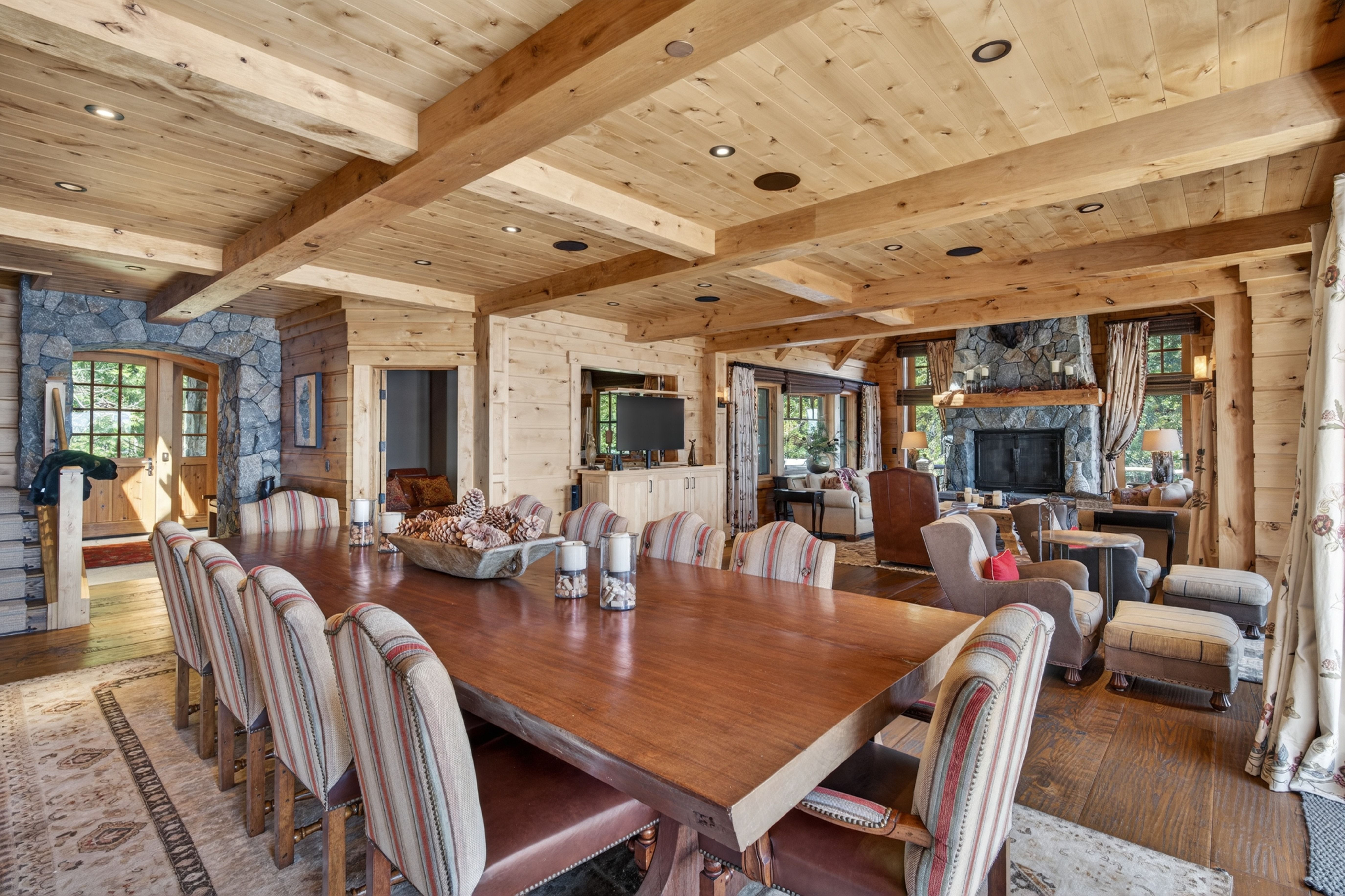 This spacious and rustic dining area features a large, rich-wood table surrounded by striped upholstered chairs, set beneath impressive vaulted ceilings with exposed log beams. The open-concept design flows seamlessly into a cozy living space anchored by a prominent stone fireplace, creating an inviting and warm mountain-lodge atmosphere. Natural textures, including wood paneling and stone accents, define the luxury interior aesthetic.