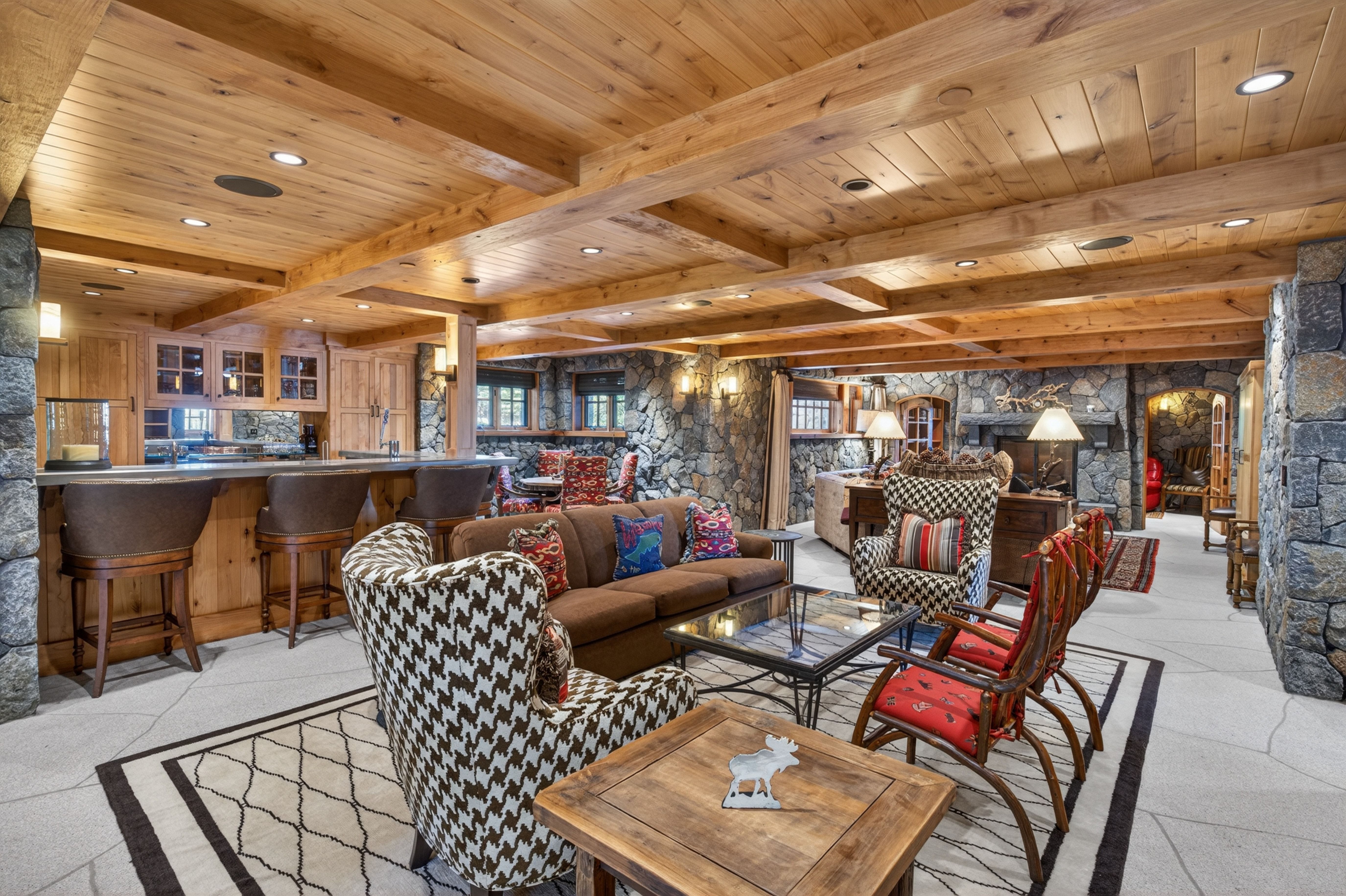 This spacious, lodge-style basement features an open-concept layout that includes a comfortable seating area, a wet bar, and a rustic stone fireplace. The room is accented by exposed wooden ceiling beams and natural stone walls, creating a cozy and luxurious cabin aesthetic. The blend of rich brown, patterned upholstery and high-end wood cabinetry makes this an ideal space for entertainment and relaxation.
