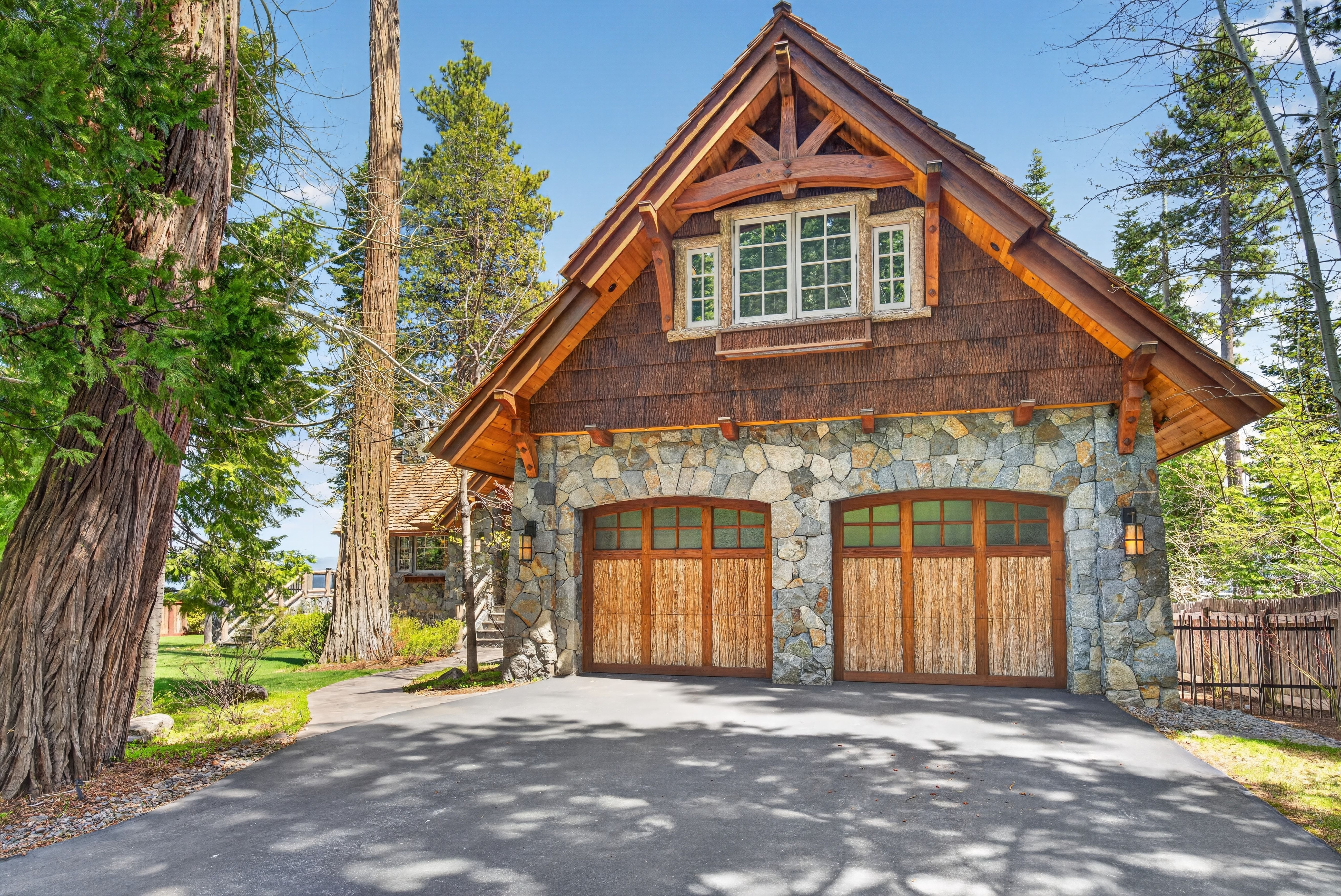 This stunning detached garage features a sophisticated rustic aesthetic, blending natural stone masonry with vertical wood shake siding and exposed timber roof trusses. The structure includes two large, custom wood carriage-style garage doors and an appealing gabled roofline. Set among towering pine trees with a well-maintained driveway, the property exudes a warm, mountain-cabin charm suitable for an upscale retreat.