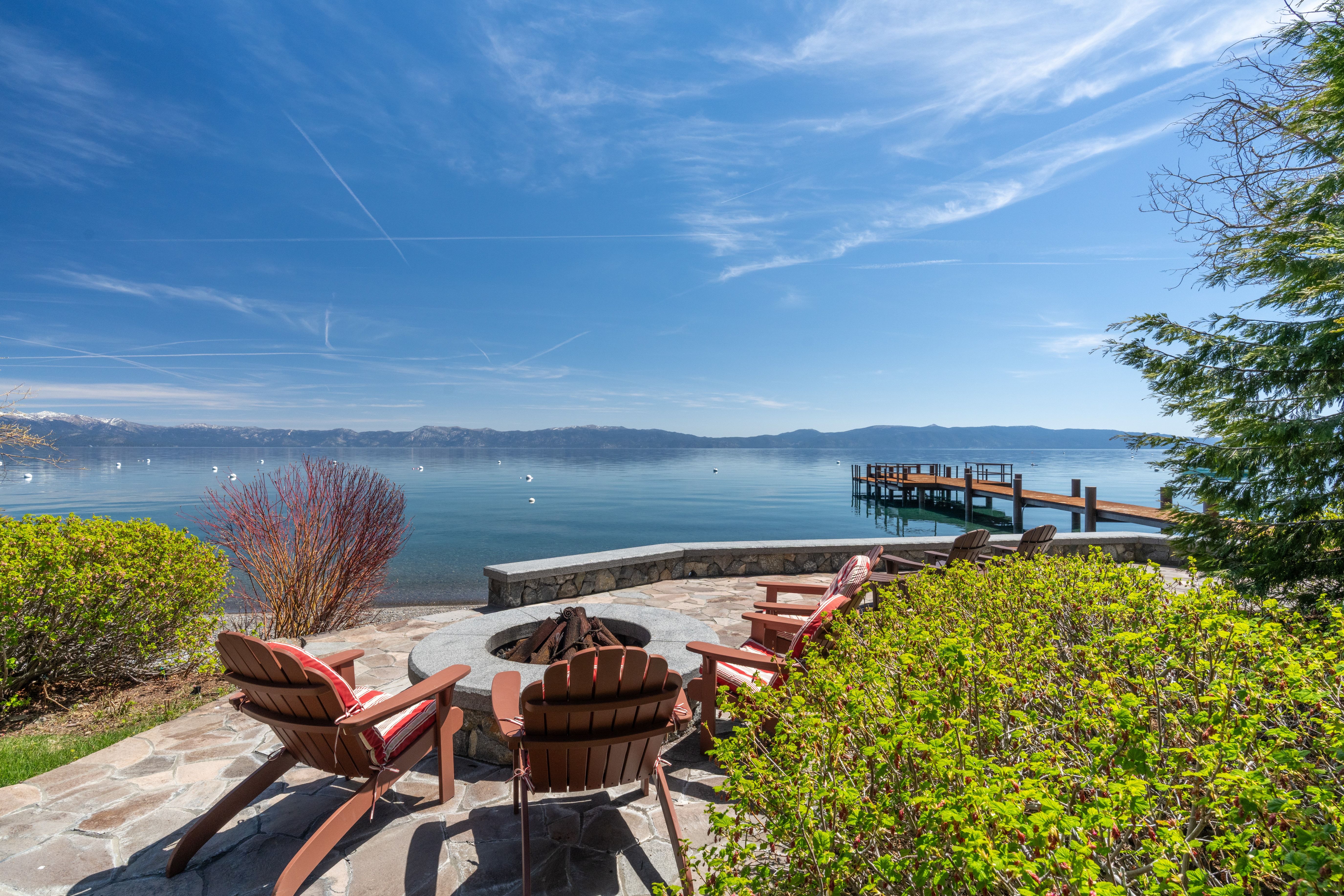 This stunning lakeside patio features a stone-paved fire pit area with Adirondack chairs, perfectly positioned for taking in panoramic water and mountain views. The space includes a private dock extending into the calm, clear lake, framed by lush landscaping and a stone retaining wall. It offers an inviting and tranquil outdoor living area ideal for waterfront entertaining or peaceful relaxation.