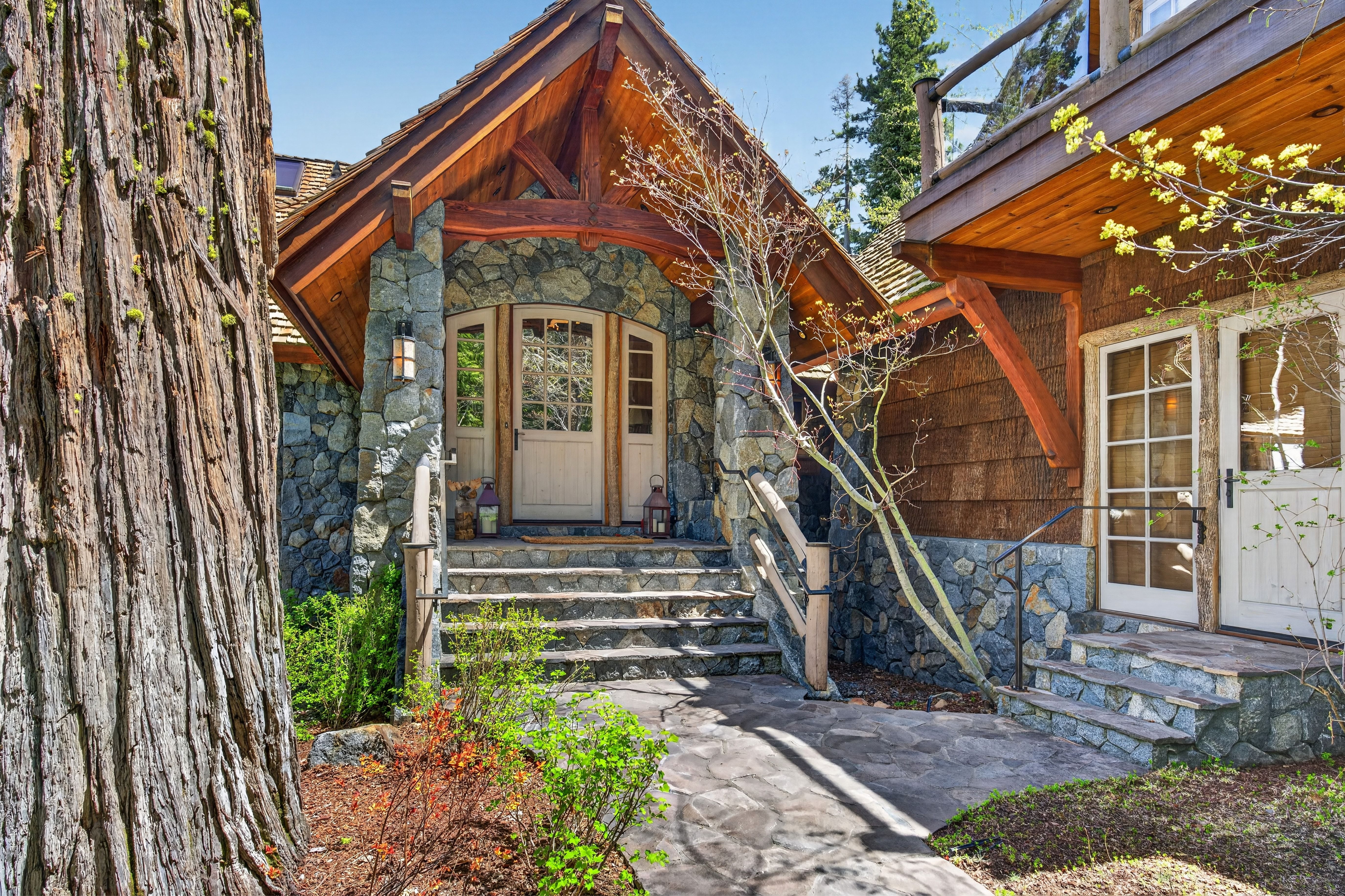 This charming mountain-style entryway features a rustic aesthetic, defined by massive, textured stone walls and warm cedar timber framing. A stone-paved walkway leads up to a set of welcoming stone steps and a grand wooden entrance door, framed by large, character-rich trees. The overall architectural design exudes a cozy, lodge-like appeal perfect for a getaway retreat.