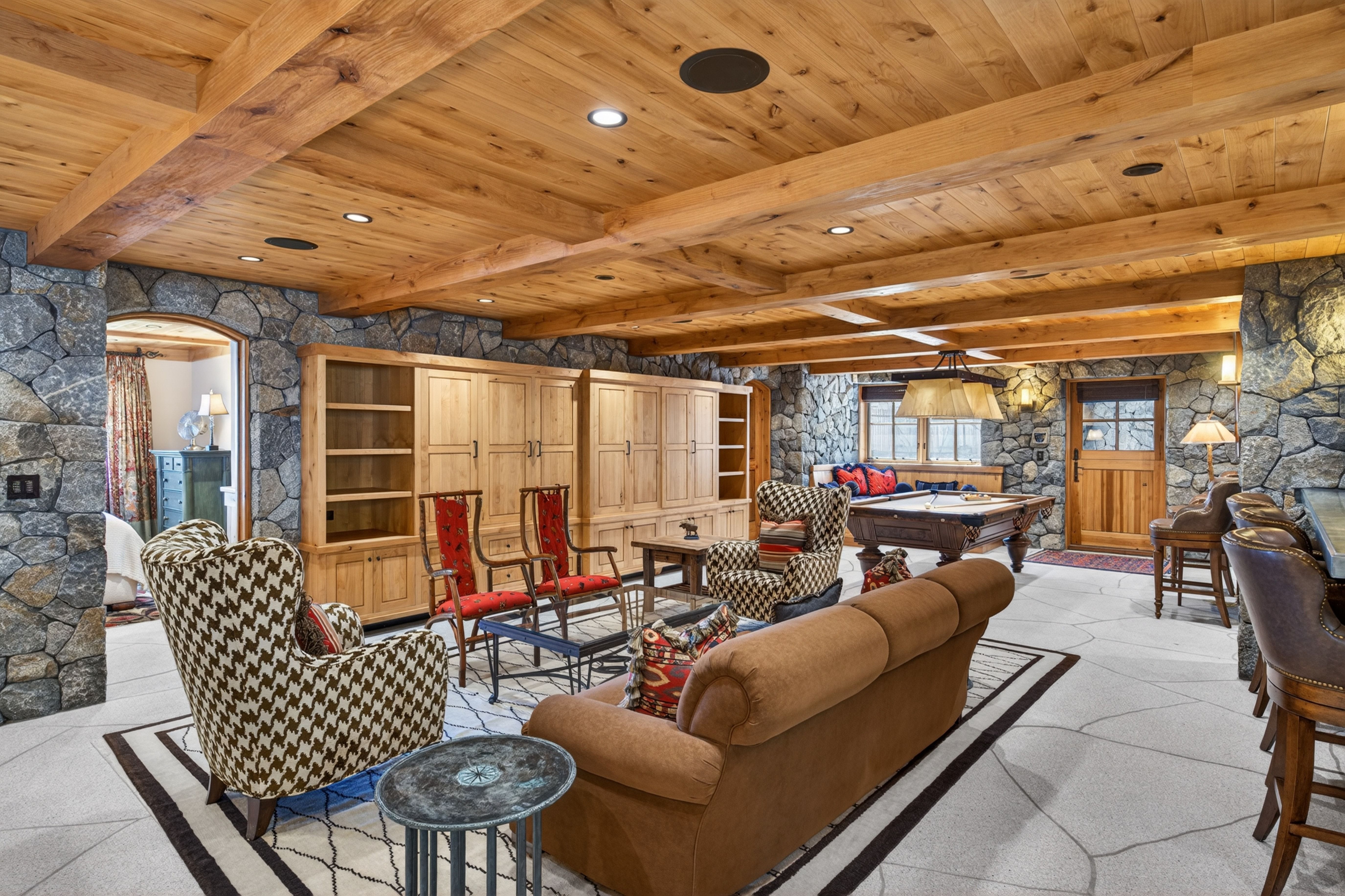 This spacious and rustic-themed finished basement features natural stone walls, warm exposed wood beam ceilings, and light-toned flagstone flooring. The room is designed for entertainment, housing a cozy living area with patterned armchairs and a comfortable sofa, as well as a billiards area, built-in cabinetry, and a bar. The large open floor plan and high-quality materials create a cohesive, lodge-inspired atmosphere perfect for hosting guests.