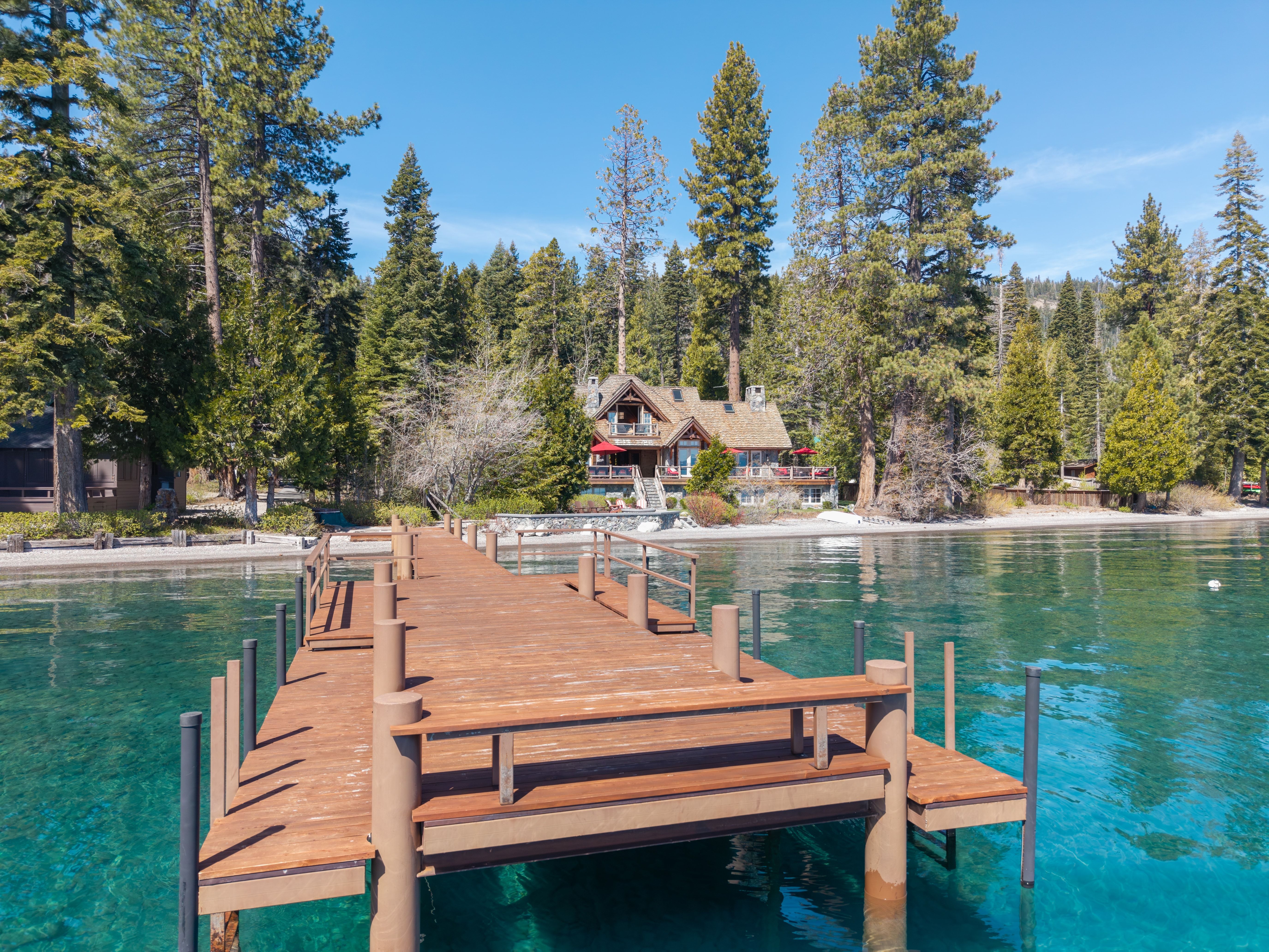 This stunning lakefront property features a classic mountain-style cabin with a spacious deck overlooking crystal-clear water. A wooden pier stretches out from the private shoreline toward the home, which is nestled amongst towering pine trees and lush greenery. The setting offers a serene, luxurious atmosphere perfect for lakefront living and outdoor recreation.