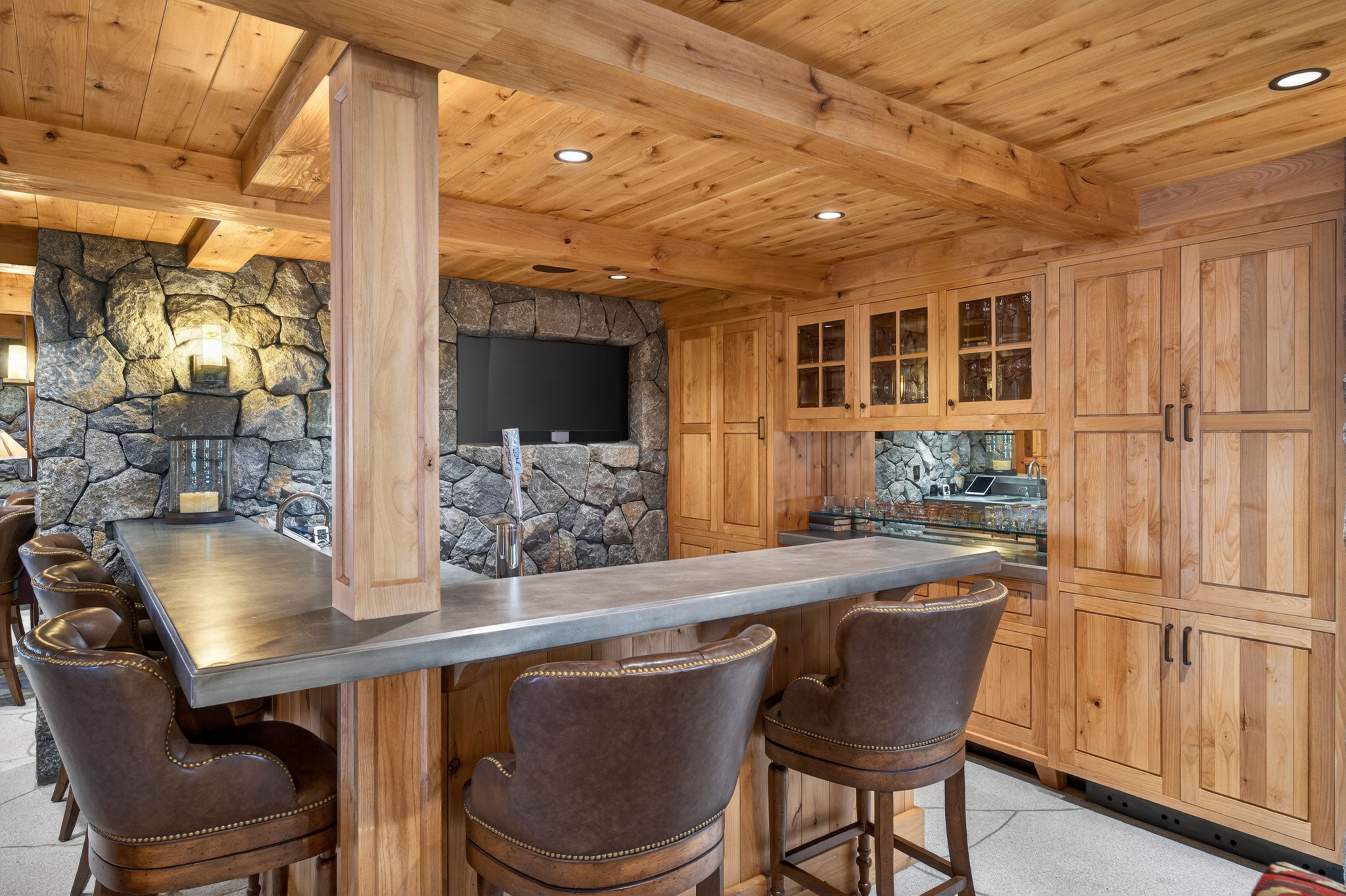 This rustic home bar area features a prominent stone accent wall, warm wood cabinetry, and a sturdy concrete-topped bar with leather barstools. The space is highlighted by integrated ceiling lighting, rich wooden beams, and a sophisticated, inviting atmosphere perfect for entertaining. It showcases high-quality craftsmanship, blending natural stone and wood textures to create a cozy, lodge-like aesthetic.