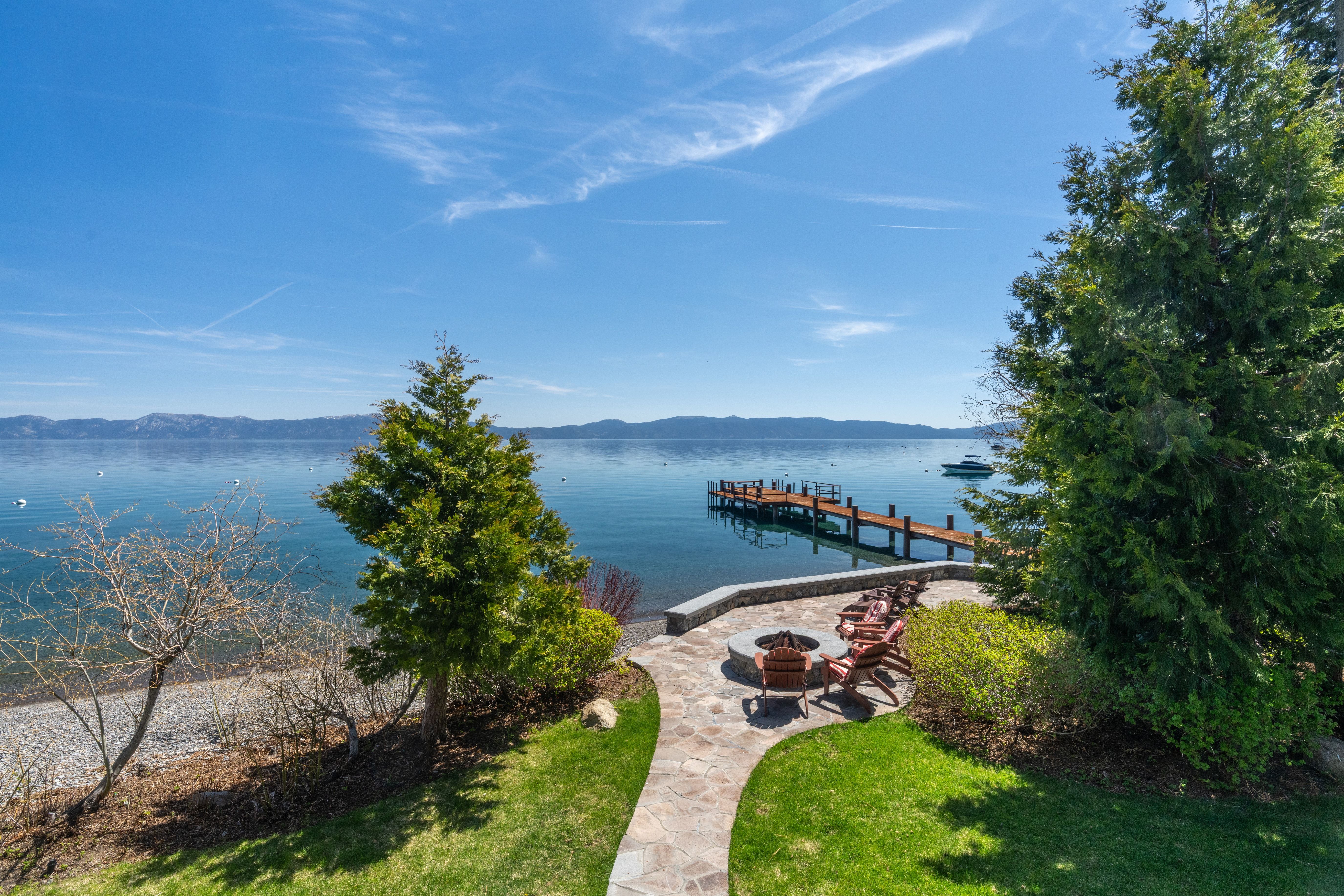 This stunning lakeside patio features natural stone paving and a built-in fire pit, offering a serene space for outdoor relaxation. Adirondack chairs are positioned to take in the panoramic water views, which include a private wooden dock extending into the calm, clear lake. The lush surrounding landscaping, mature trees, and mountainous backdrop create an inviting and peaceful atmosphere ideal for summer entertaining.