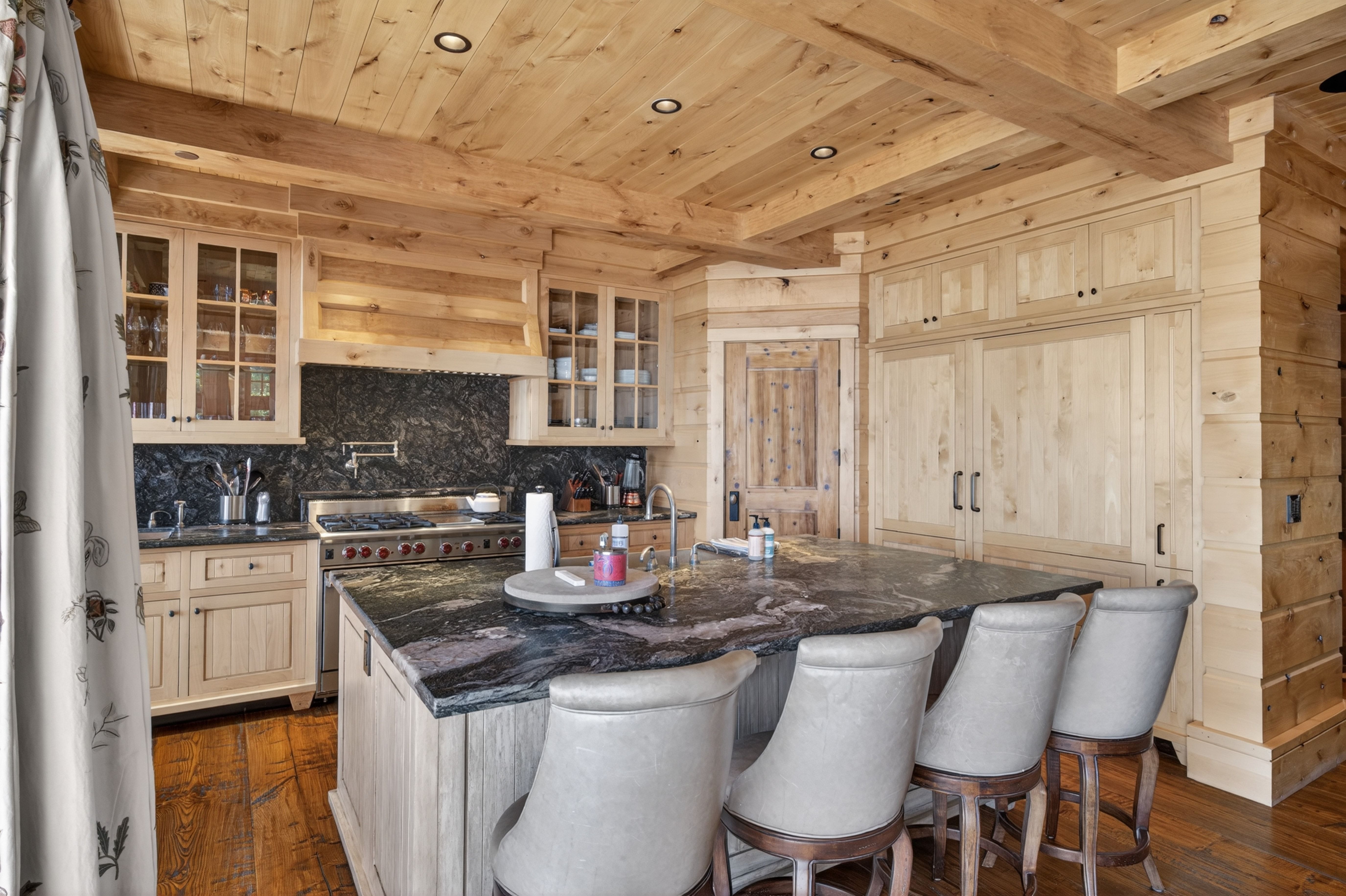 This rustic, high-end kitchen features natural wood paneling and cabinetry throughout, paired with a commanding black granite countertop on the large center island. The space is well-equipped with professional-grade stainless steel appliances, a pot filler, and four elegant leather-upholstered bar stools. The warm wood tones and rich stone textures create a cozy yet sophisticated atmosphere perfect for both cooking and entertaining.