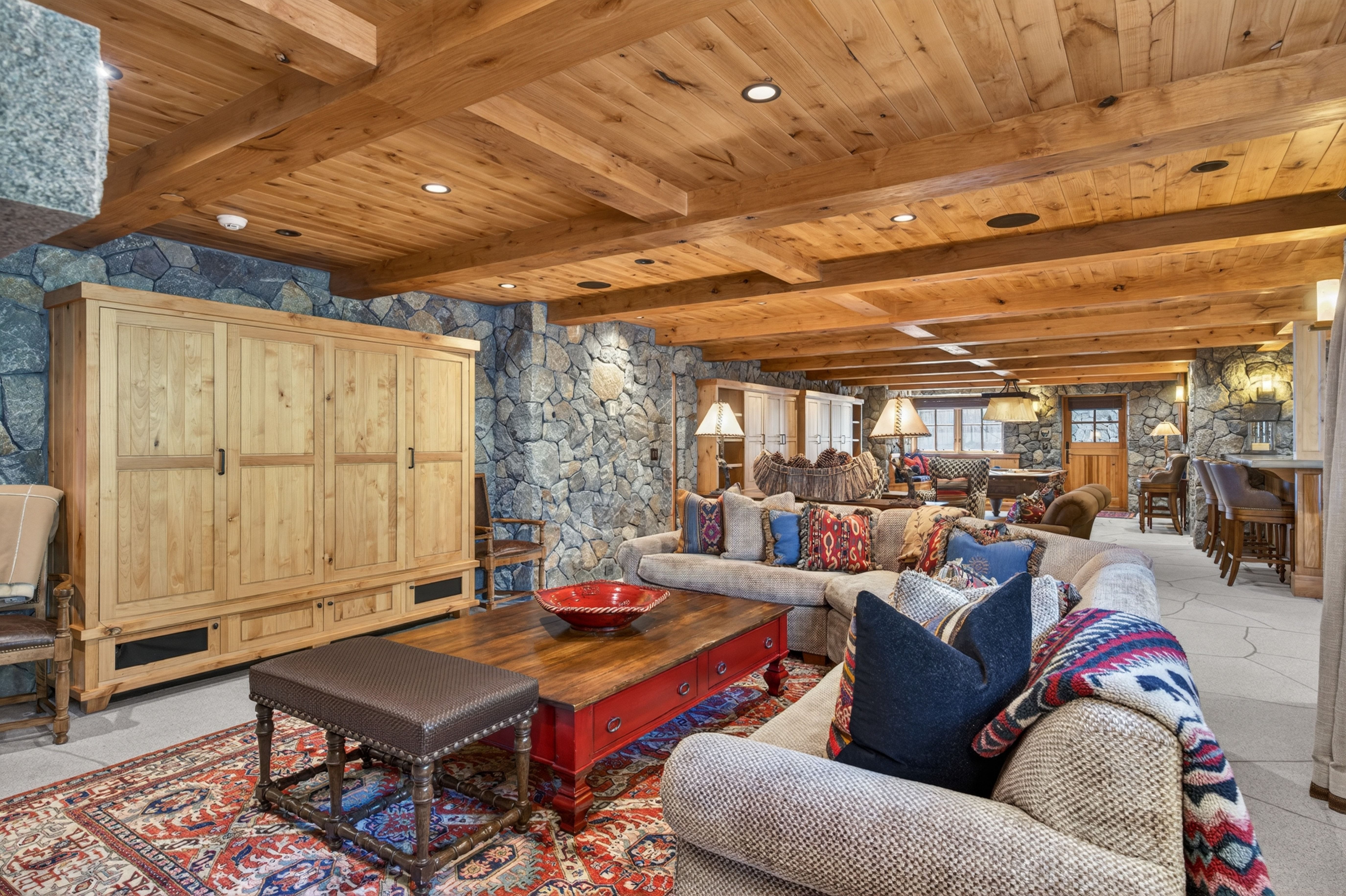 This spacious, open-concept living area features a warm rustic aesthetic characterized by heavy wooden beam ceilings and textured stone wall accents. A comfortable, sprawling sectional sofa is adorned with patterned and solid throw pillows, centered around a large red coffee table with an upholstered ottoman. The room is further accented by an ornate Persian-style area rug, recessed lighting, and a seamless flow into a secondary lounge space with a game table and wet bar area.