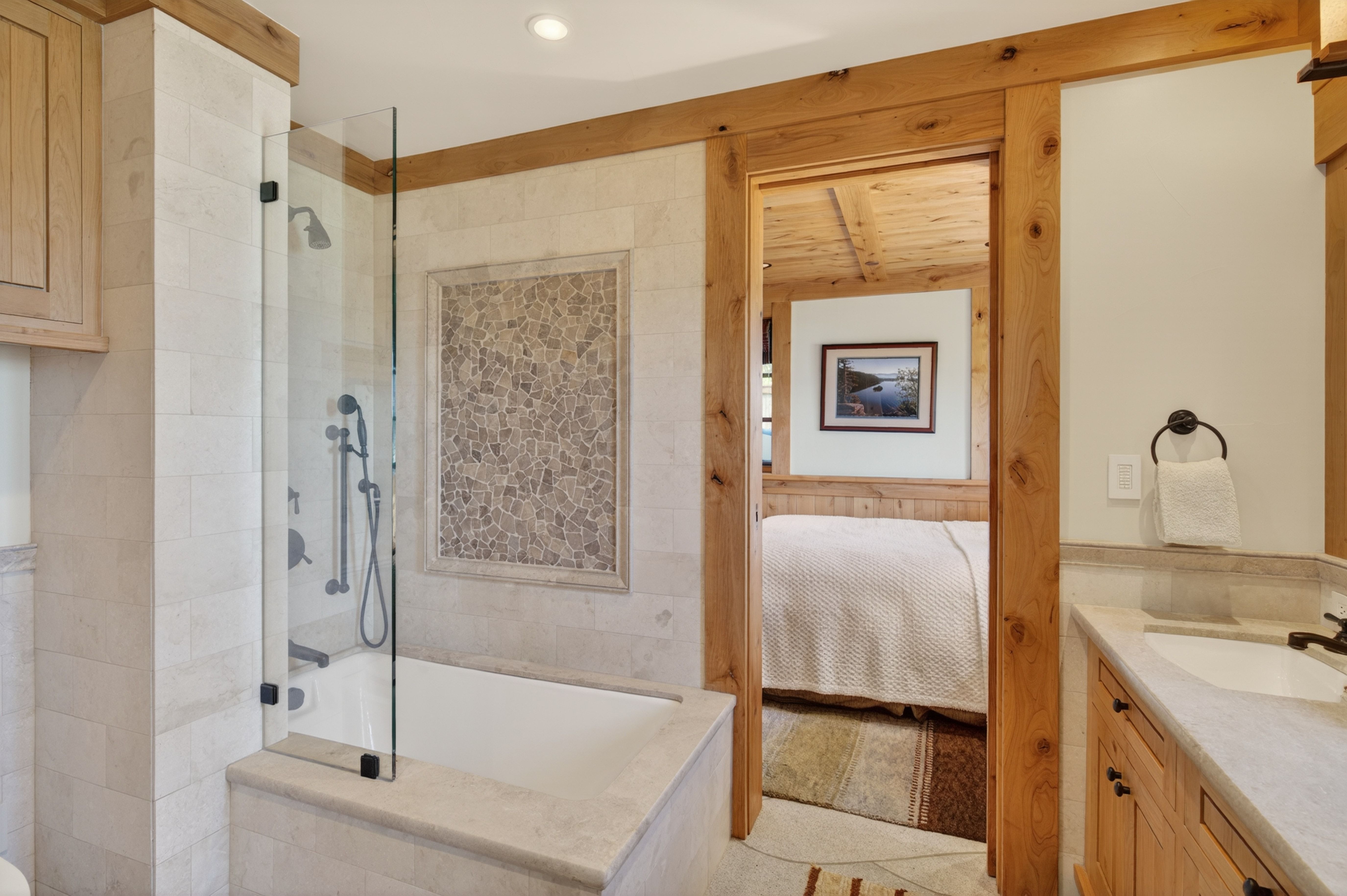 This primary bathroom features a rustic, lodge-inspired aesthetic with warm wood trim and natural stone accents. It includes a built-in tub with an integrated shower enclosure, a stone-topped vanity, and a view into a cozy adjacent bedroom. The use of neutral colors and rich textures provides a sophisticated yet comfortable atmosphere.