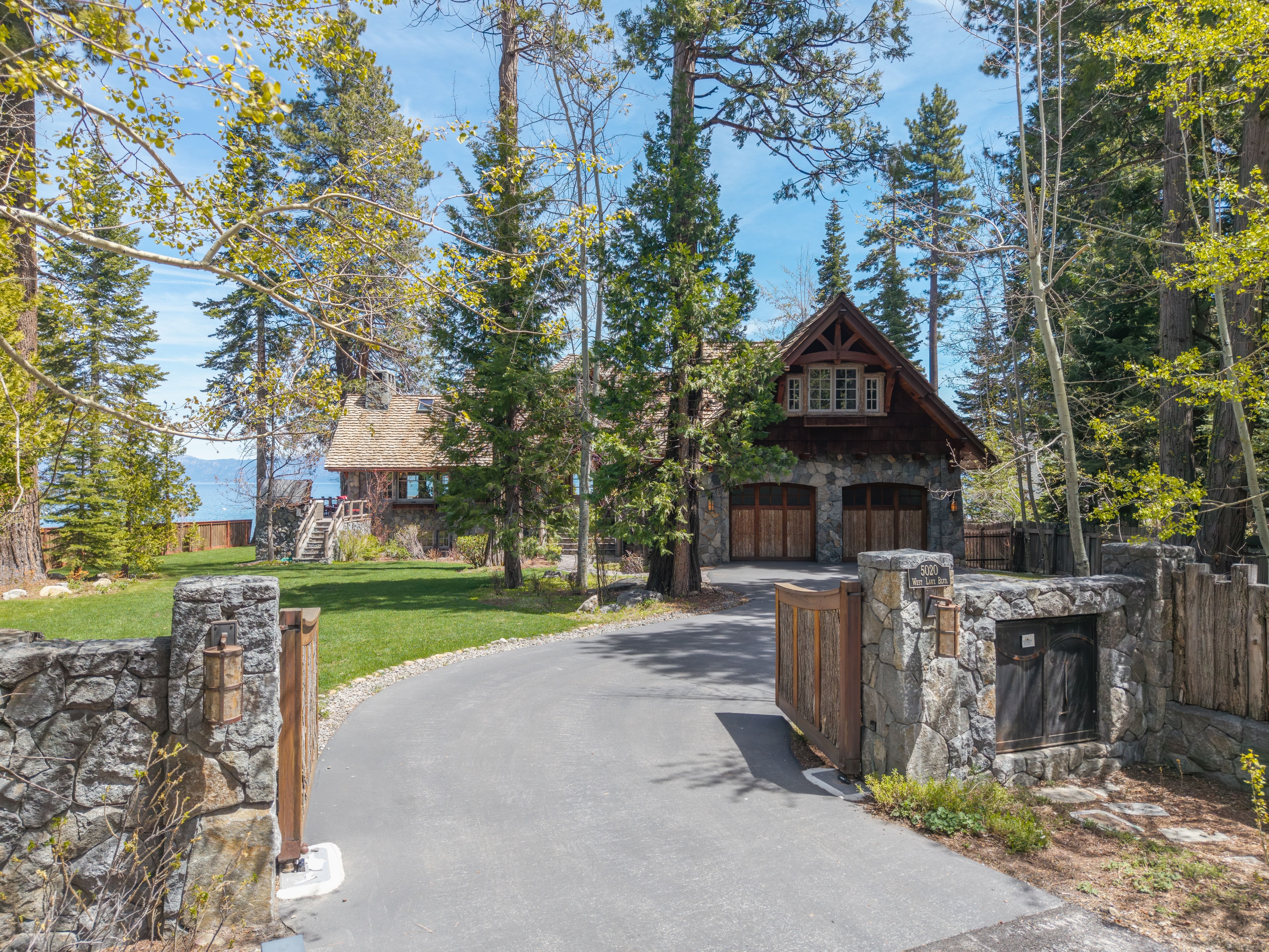 This elegant mountain-style residence showcases a beautiful combination of natural stone masonry and timber wood accents, nestled within a lush, forested landscape. A sweeping paved driveway leads through a private gated entrance to an attached two-car garage, with the property offering scenic views of the water in the background. The home's design exudes a rustic yet sophisticated charm, well-integrated into its serene natural surroundings.