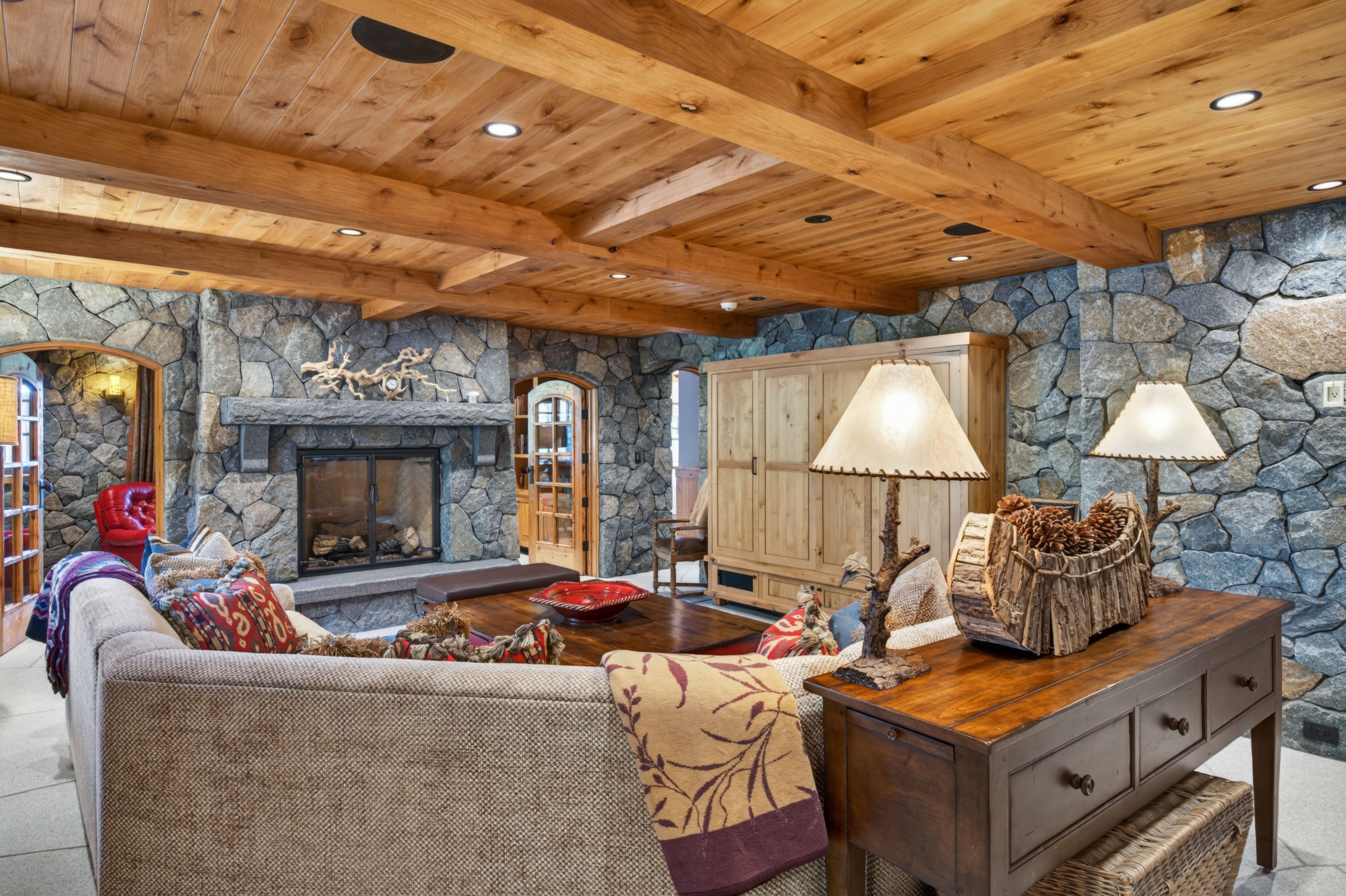 This cozy living room features striking stone walls, rustic exposed beam ceilings, and a large fireplace that anchors the space with warmth and character. The room is furnished with a comfortable sofa and a rustic wooden console table, enhanced by decorative cabin-style accents. The overall aesthetic is rustic, warm, and inviting, showcasing high-quality woodwork and natural elements.