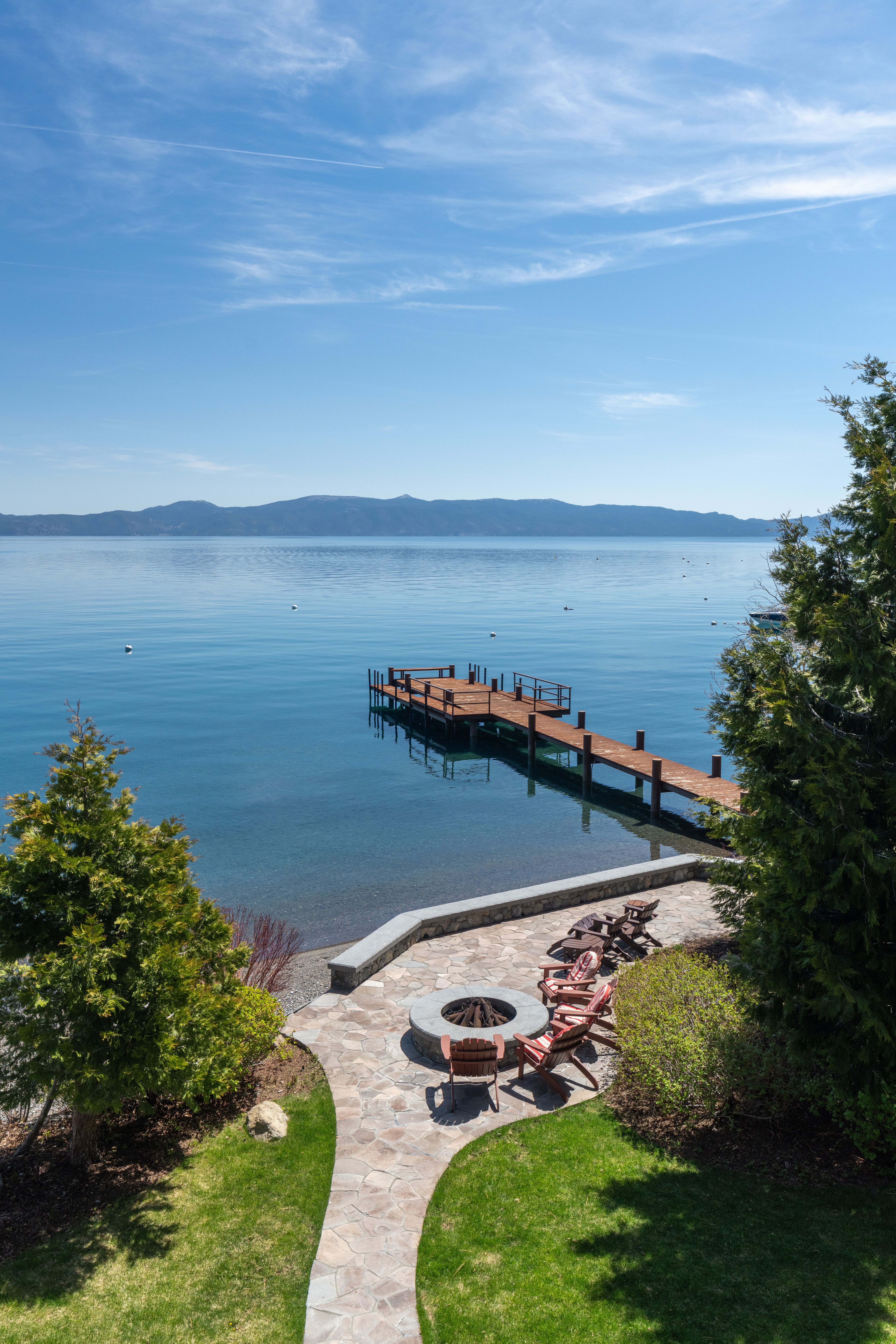 This serene lakefront patio features a stone-paved seating area arranged around a circular fire pit, offering an ideal space for outdoor relaxation and entertaining. The patio overlooks a long, private wooden dock extending into the calm, deep blue water of the lake, with picturesque mountains visible in the distance. The combination of lush greenery, stone masonry, and expansive water views creates a tranquil, high-end lakeside retreat.
