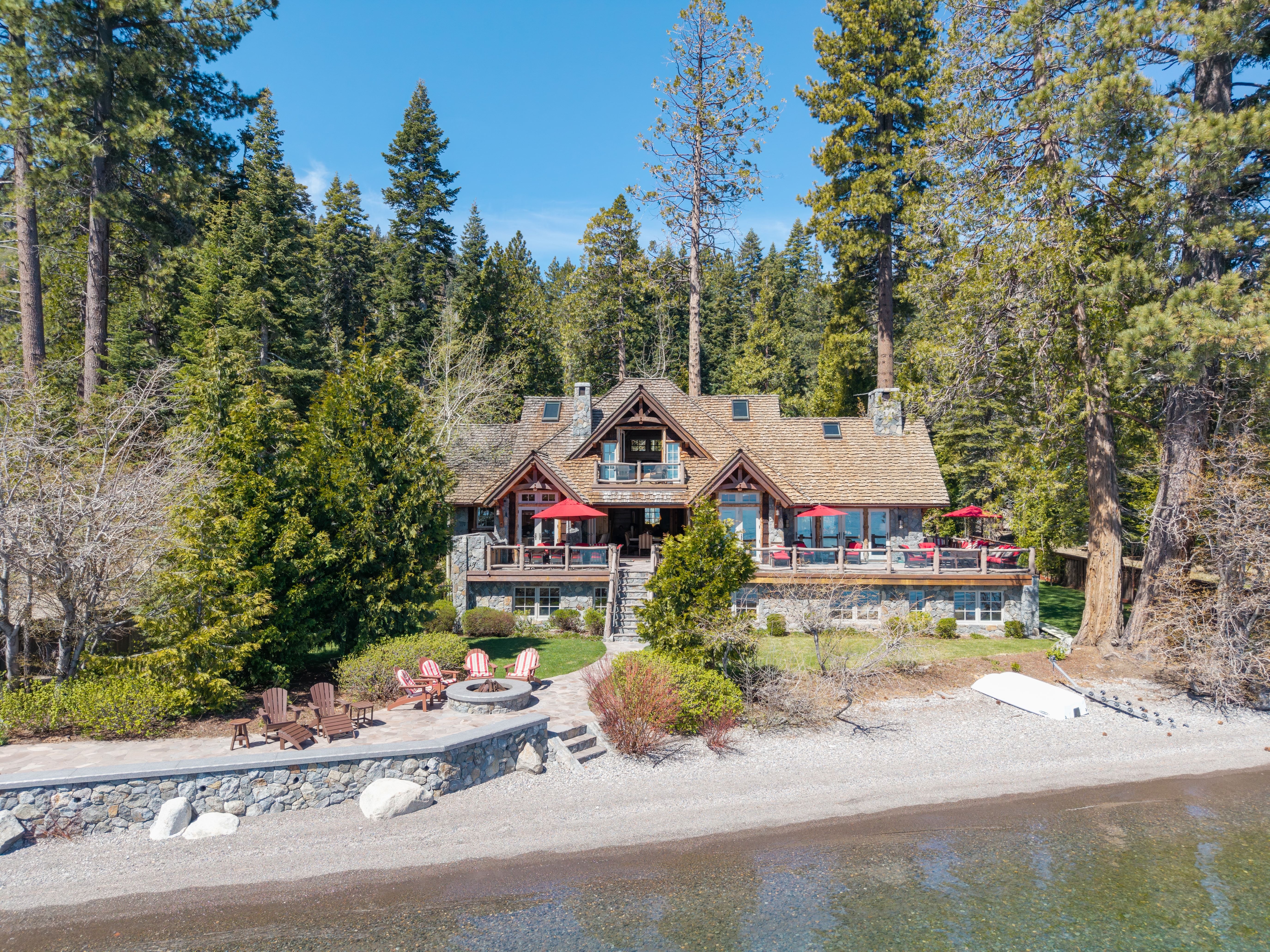 This scenic lakefront estate showcases a sophisticated mountain-style architecture with extensive stone masonry and timber framing. The property features a multi-level deck overlooking the water, complete with red-accented seating areas, a private stone patio with a fire pit, and direct beach access. Nestled within a lush, wooded landscape, this residence offers an idyllic and serene retreat.