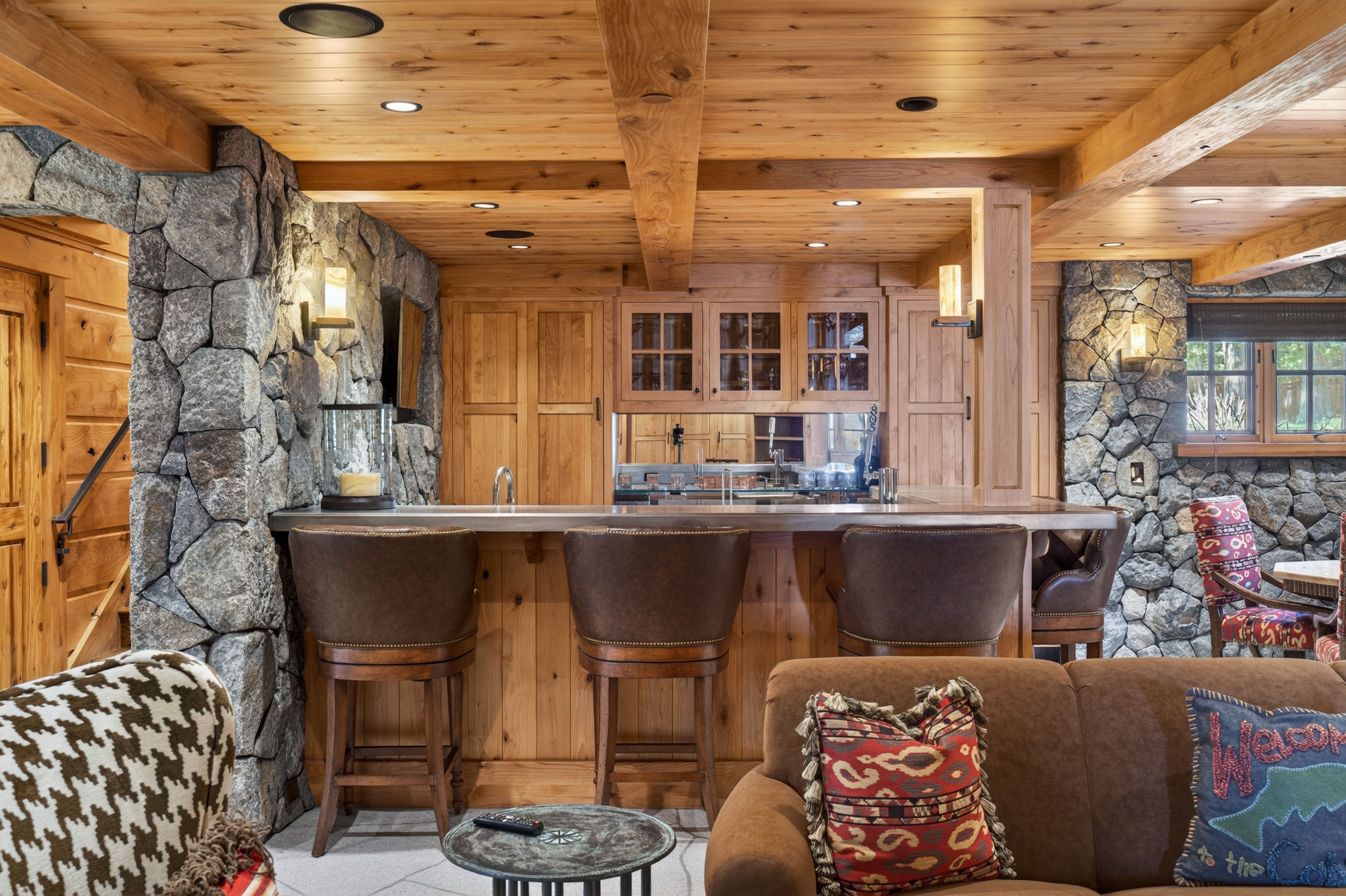 This cozy, rustic-style living area features a built-in wet bar with dark leather stools, natural wood ceiling beams, and distinctive stone-clad walls. The space is warm and inviting, anchored by a brown sofa with decorative patterned pillows and a small metallic side table. It skillfully blends rugged, mountainous architecture with comfortable, traditional furnishings, making it an ideal space for entertaining or relaxing.