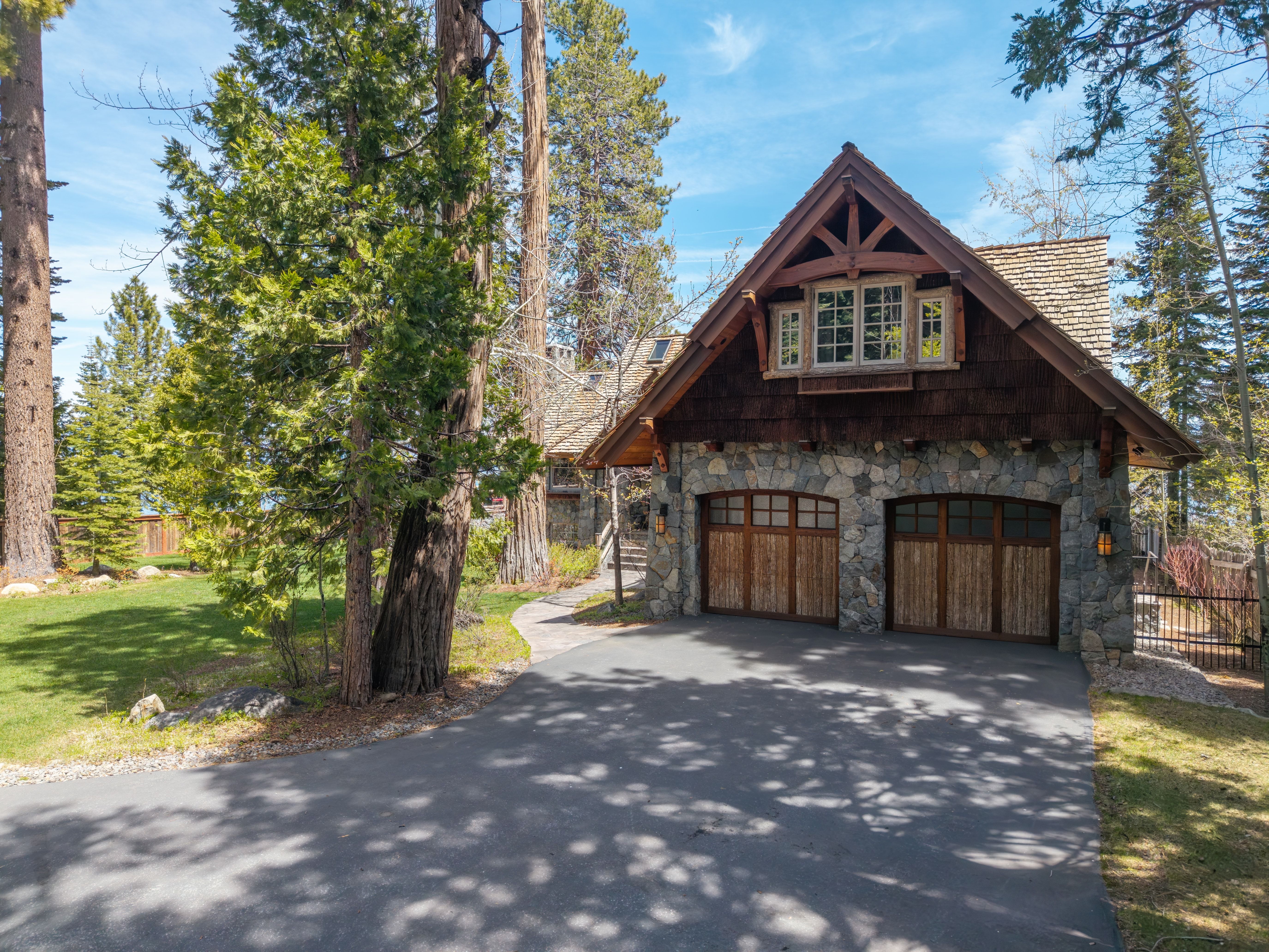 This stunning mountain-style garage features natural stone masonry, cedar shingle siding, and detailed timber framing. The double-car garage includes high-quality wood doors and a charming upper-level dormer window, all situated in a picturesque, wooded landscape. The home's rustic aesthetic is perfectly complemented by the lush surroundings and asphalt driveway.