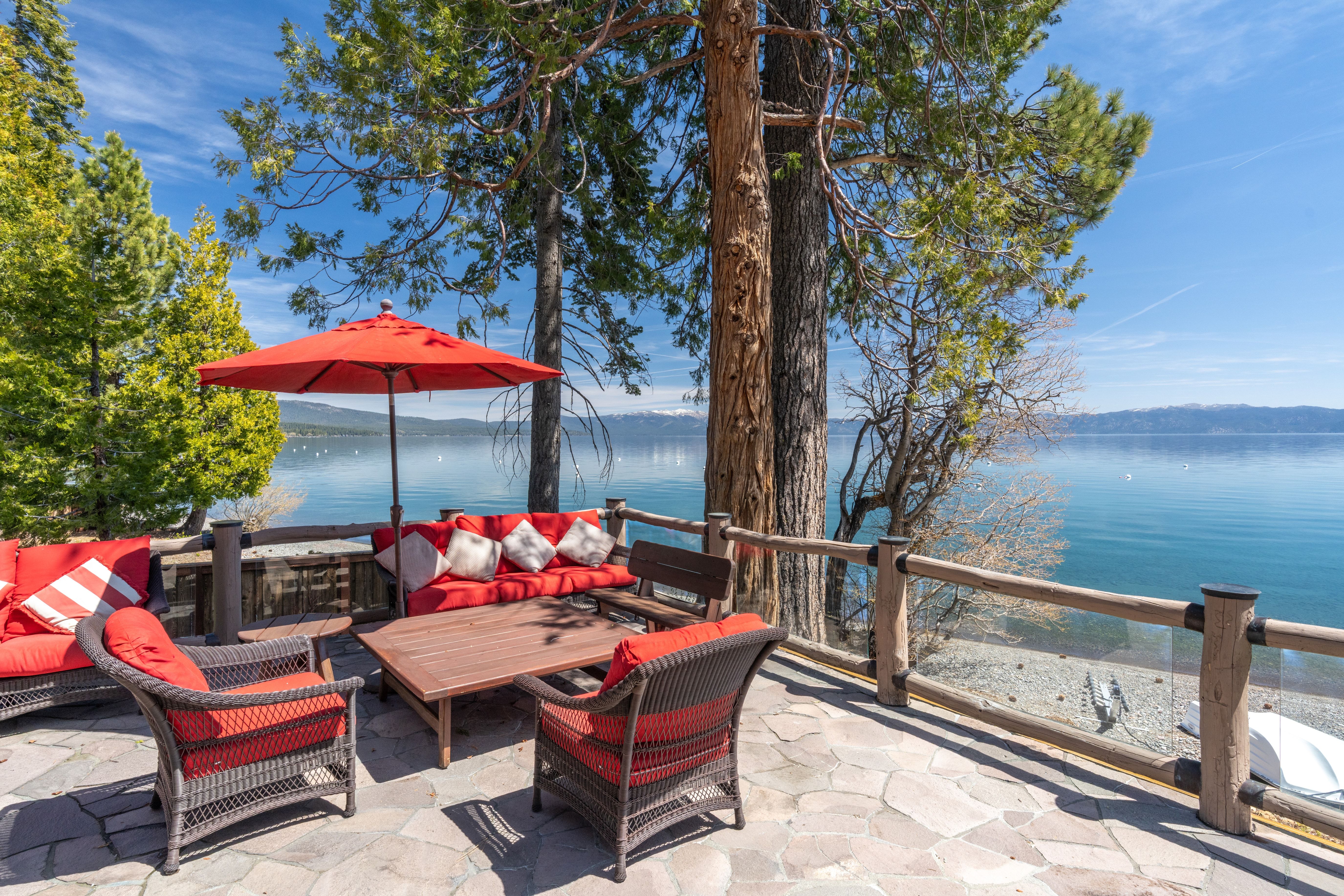 This scenic stone patio deck offers an unparalleled relaxing space overlooking a tranquil blue lake framed by majestic pine trees and distant, snow-capped mountains. The area is furnished with comfortable outdoor lounge seating, vibrant red cushions, and a large shade umbrella, creating an inviting atmosphere for outdoor entertaining or peaceful reflection. The setting exudes a luxurious, peaceful lifestyle characteristic of high-end lakeside homes.