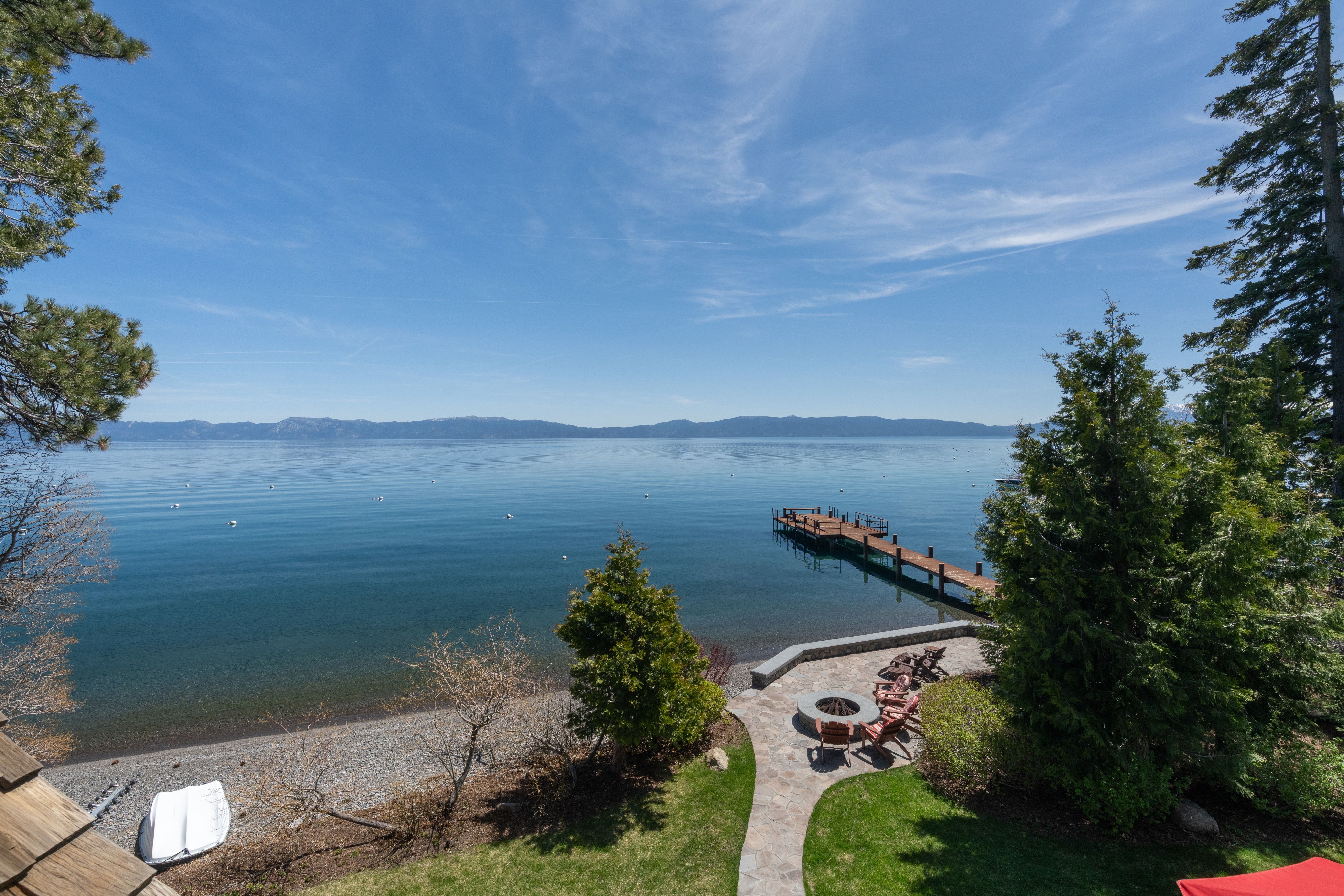 This scenic rear view captures a stunning lakeside property featuring a stone patio with a built-in fire pit and Adirondack chairs overlooking the water. A private wooden pier extends from the pebbled shoreline into the vast blue lake, which is framed by lush evergreen trees and distant mountain ranges. The tranquil setting offers an exceptional outdoor living space ideal for relaxation and panoramic views.