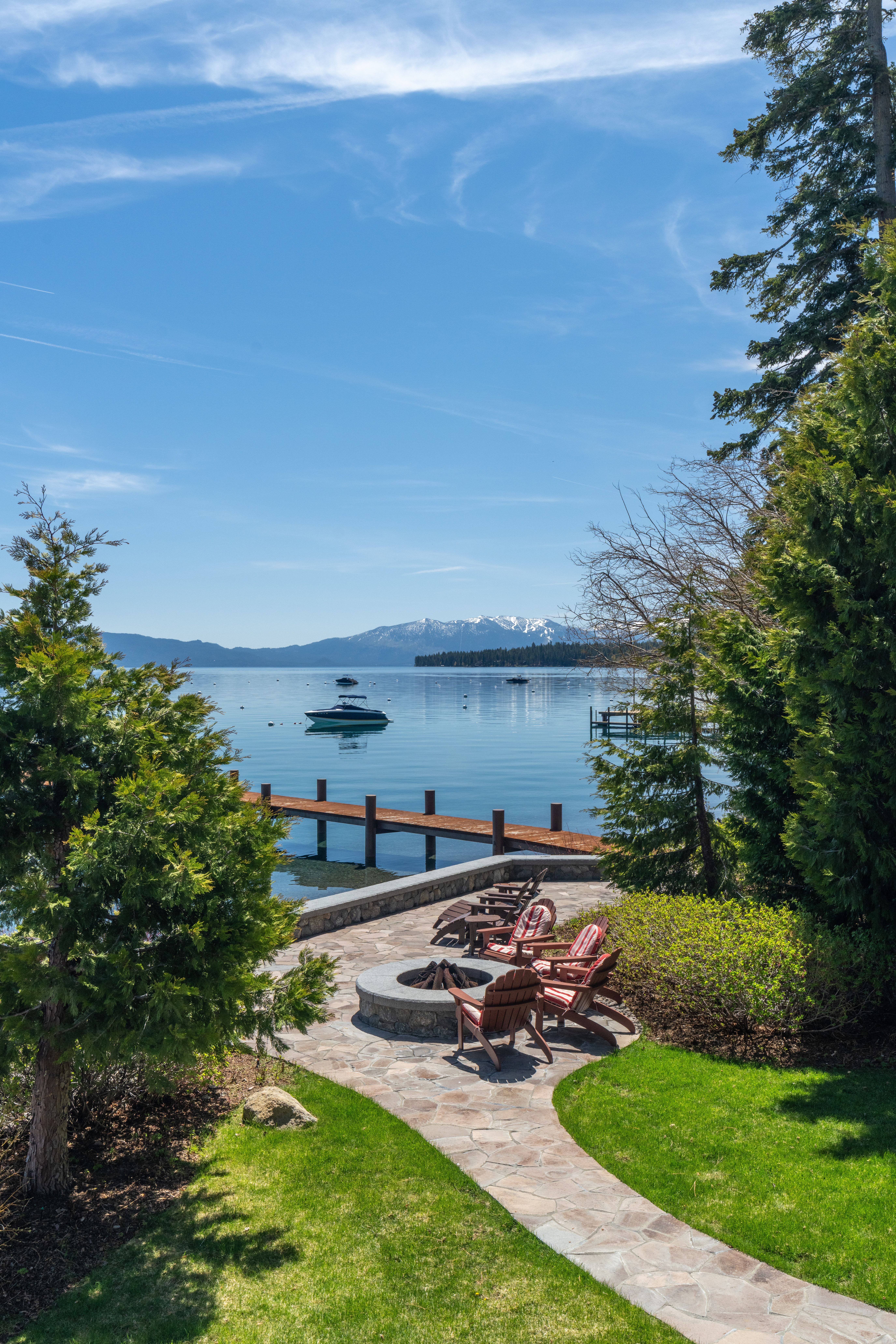 This tranquil outdoor patio features natural stone paving and a built-in fire pit, offering an ideal setting for relaxation. Surrounded by lush greenery and framed by mature trees, the space provides stunning, direct views of the lake, distant snow-capped mountains, and a private dock. Its scenic waterfront location makes it a highly desirable amenity for lakefront living.