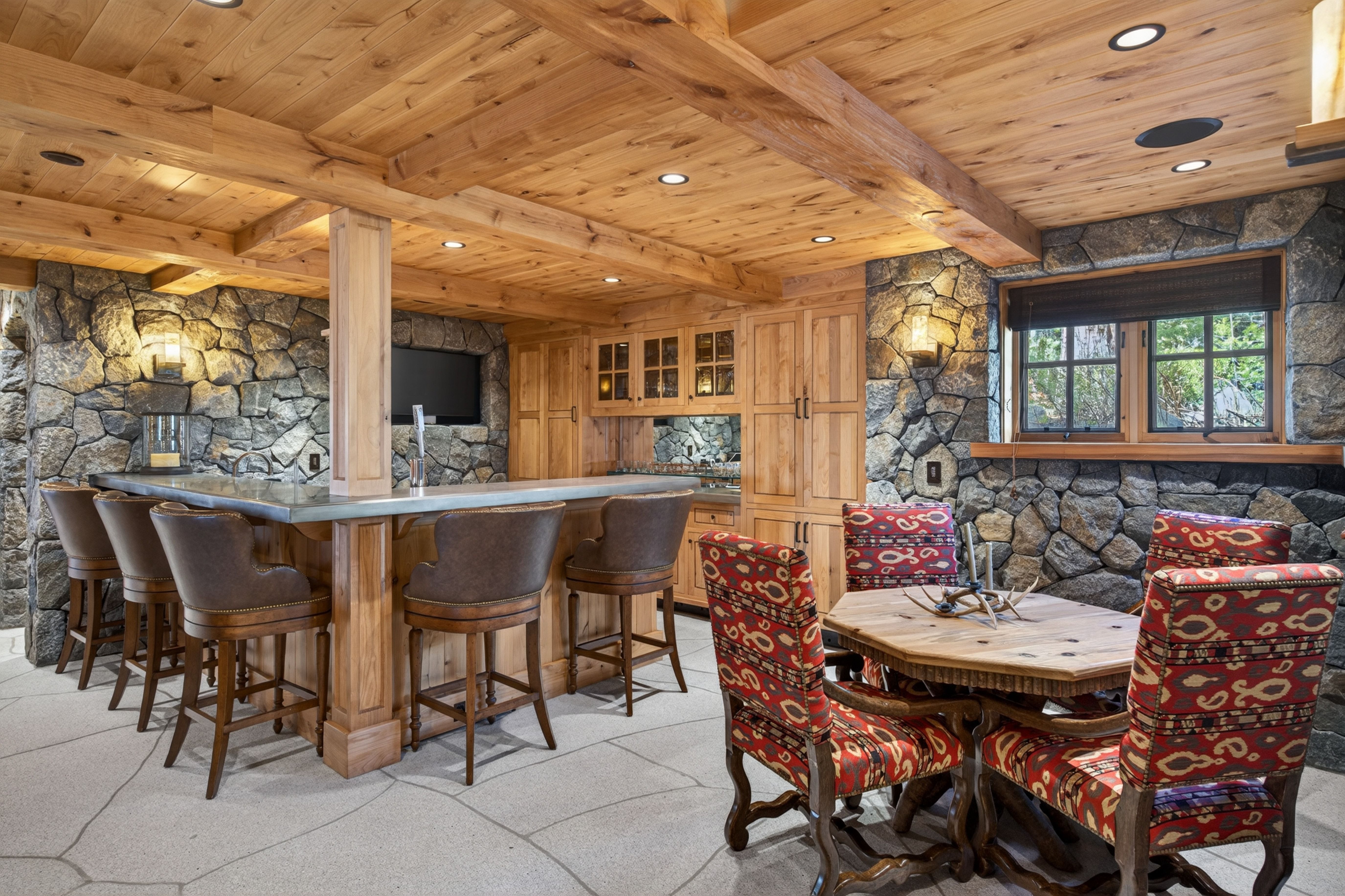 This rustic, lodge-style basement features a full bar area with leather stools, a wooden ceiling, and dramatic stone wall accents. A cozy dining nook with patterned chairs and a wooden table adds additional functionality to the space. The stone flooring and exposed wood beams create a warm, inviting atmosphere perfect for entertaining.