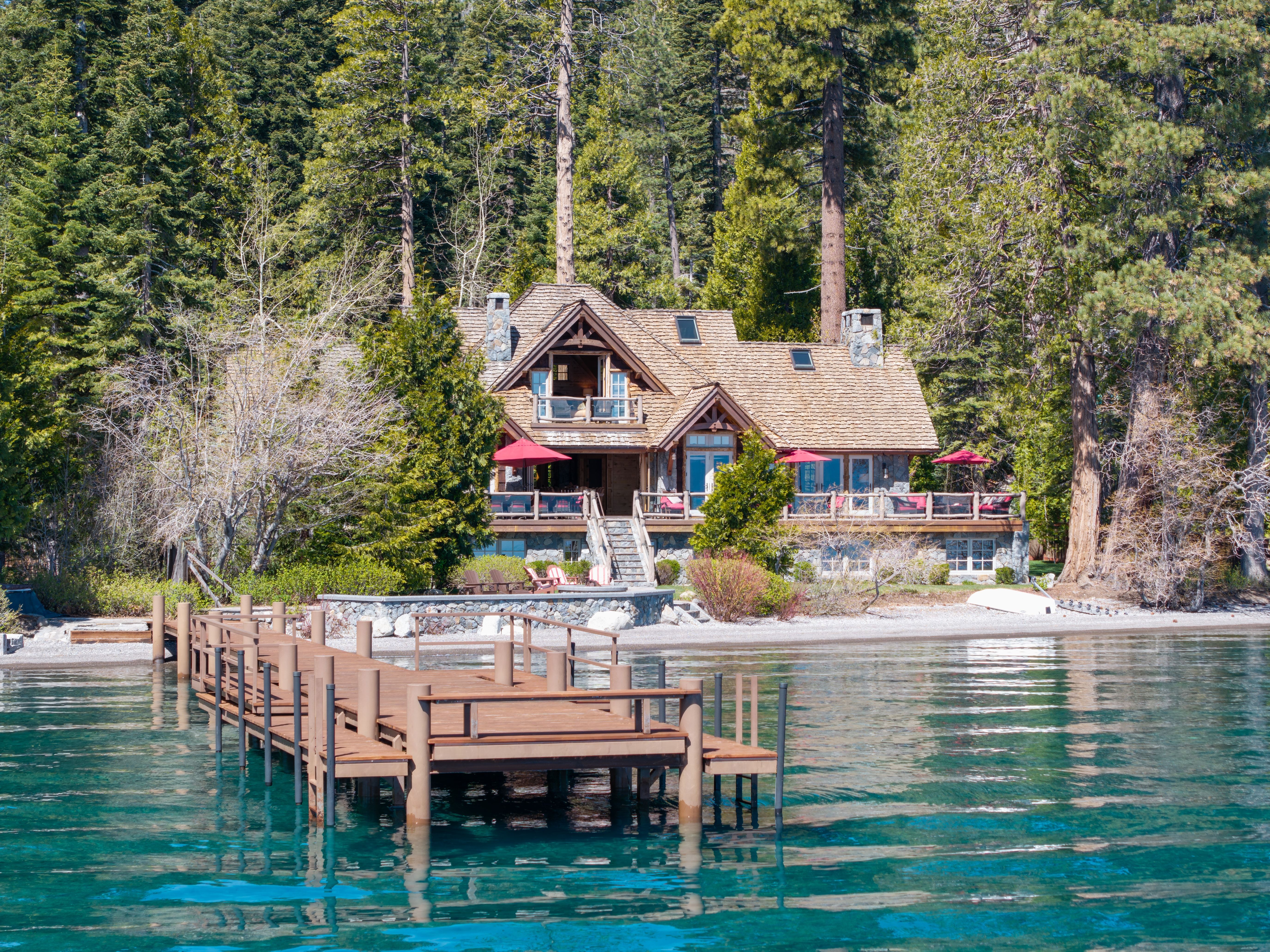 This scenic lakefront property showcases a charming, rustic-style home nestled amongst lush evergreen trees, featuring authentic stone masonry and wood accents. The expansive waterfront deck, complete with red umbrellas, provides an inviting space for relaxation while overlooking a private wooden dock extending into the clear, tranquil waters. Its serene setting and prime location make it a quintessential luxury mountain retreat.