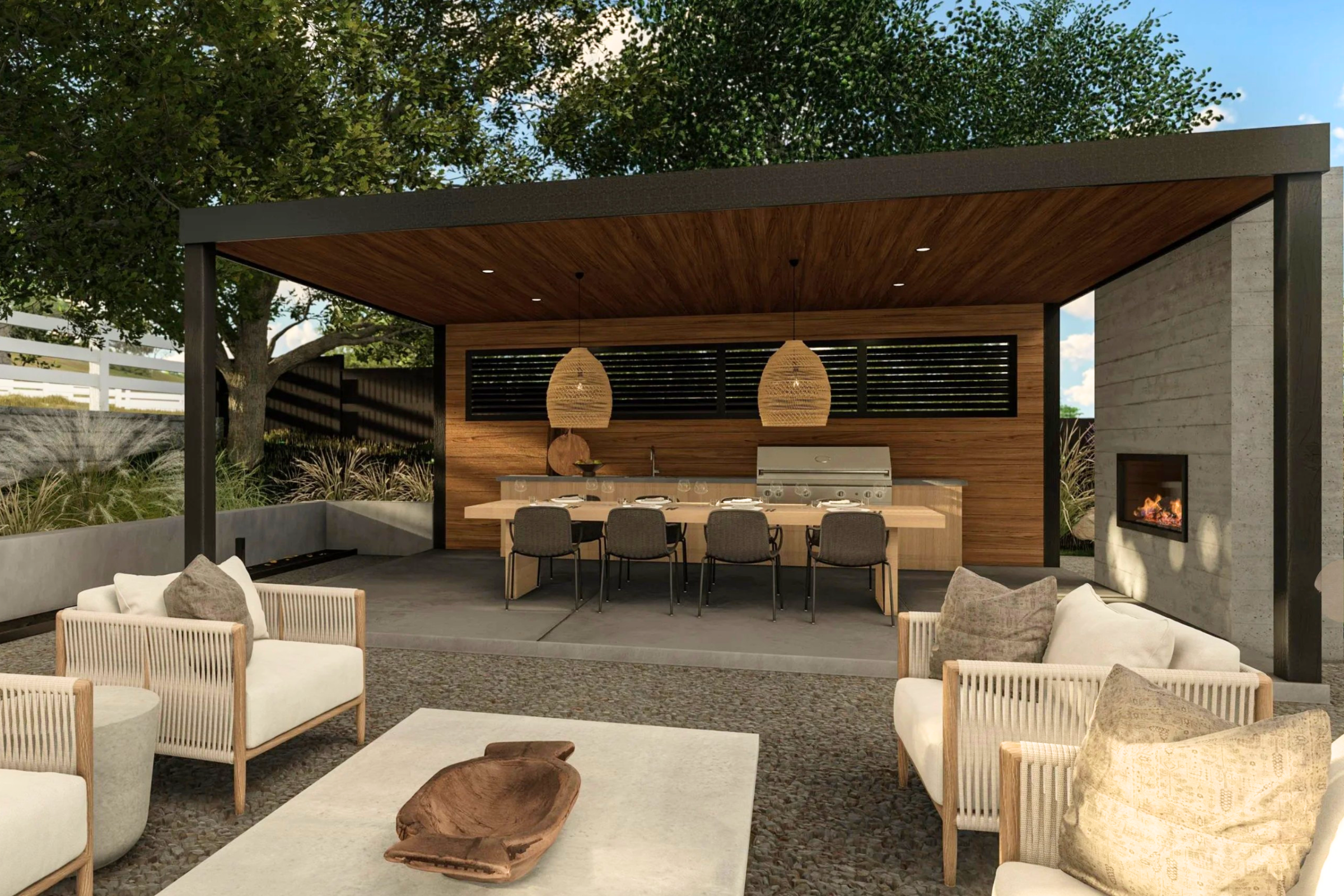 This sophisticated outdoor living space features a covered patio area with a built-in kitchen, dining table, and a cozy fireplace integrated into a concrete wall. The design blends modern architectural elements with natural wood textures and comfortable lounge seating, creating an inviting atmosphere for entertaining. The perspective is a wide, eye-level shot that captures the seamless transition between the dining area and the relaxed outdoor lounge.