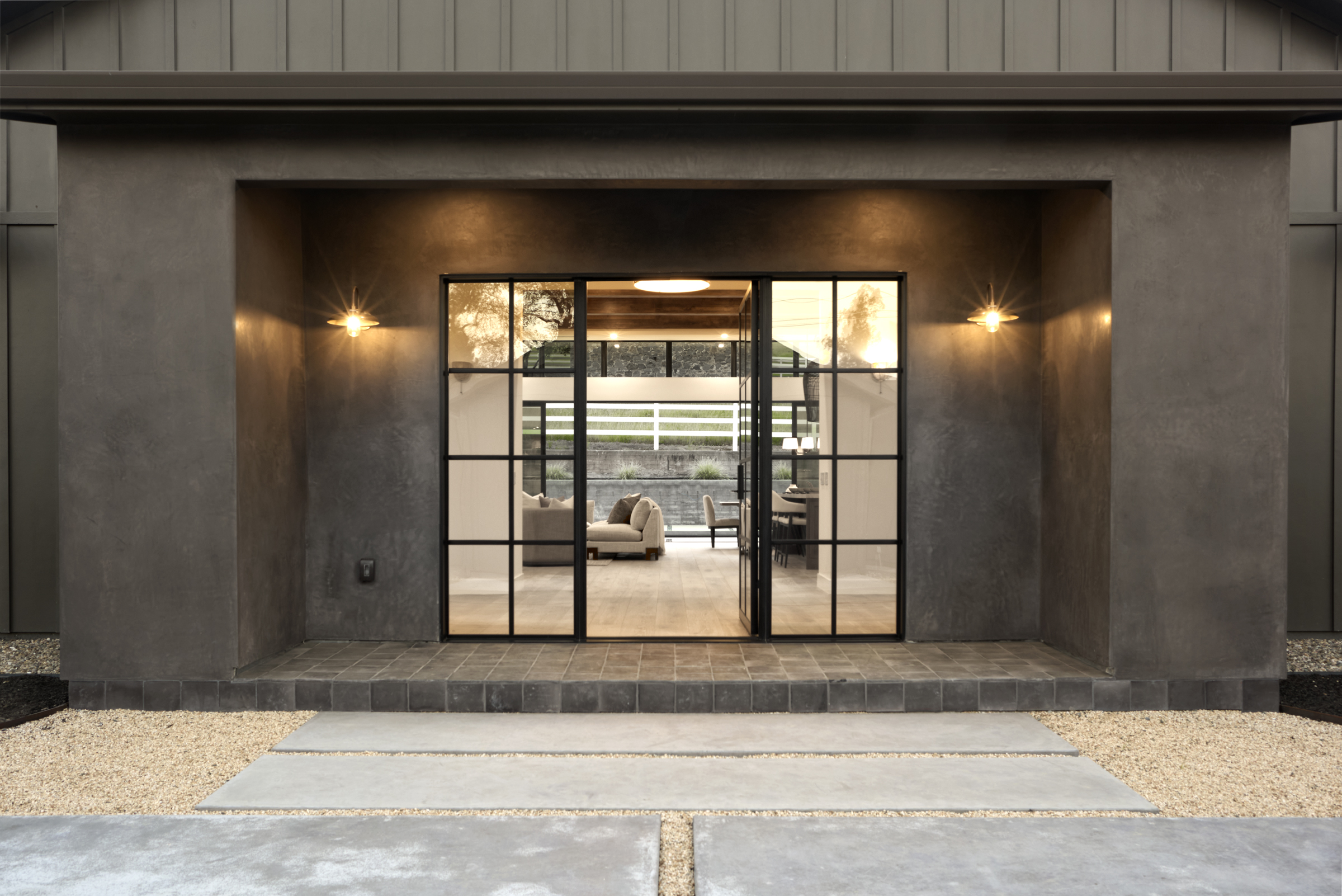 This striking entryway features a modern, dark-toned facade with a recessed porch and large, black-framed glass doors that offer a clear view into the bright, open-concept interior. The transition from the gravel and concrete walkway to the tiled porch creates a sophisticated, high-contrast aesthetic. The warm glow from the wall-mounted sconces adds an inviting touch to the minimalist, contemporary architectural design.
