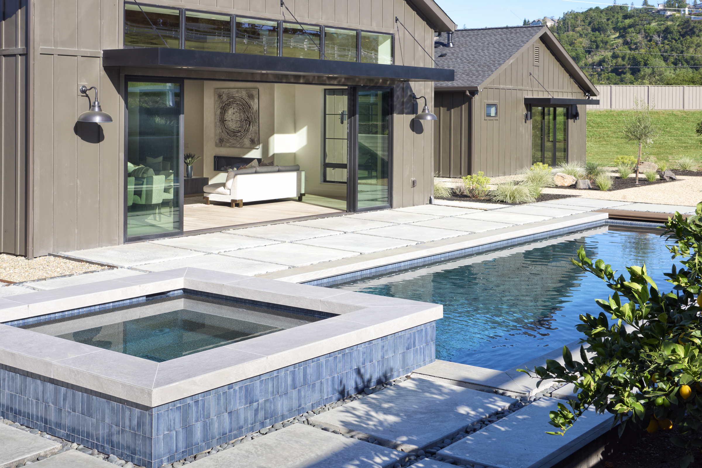 This stunning outdoor living space features a modern, dark-toned farmhouse-style home with large sliding glass doors that open to a spacious concrete patio. In the foreground, a sleek, tiled spa flows into a rectangular swimming pool, creating a seamless transition between the indoor and outdoor areas. The scene is bathed in bright, natural sunlight, highlighting the clean lines and contemporary design of the property.