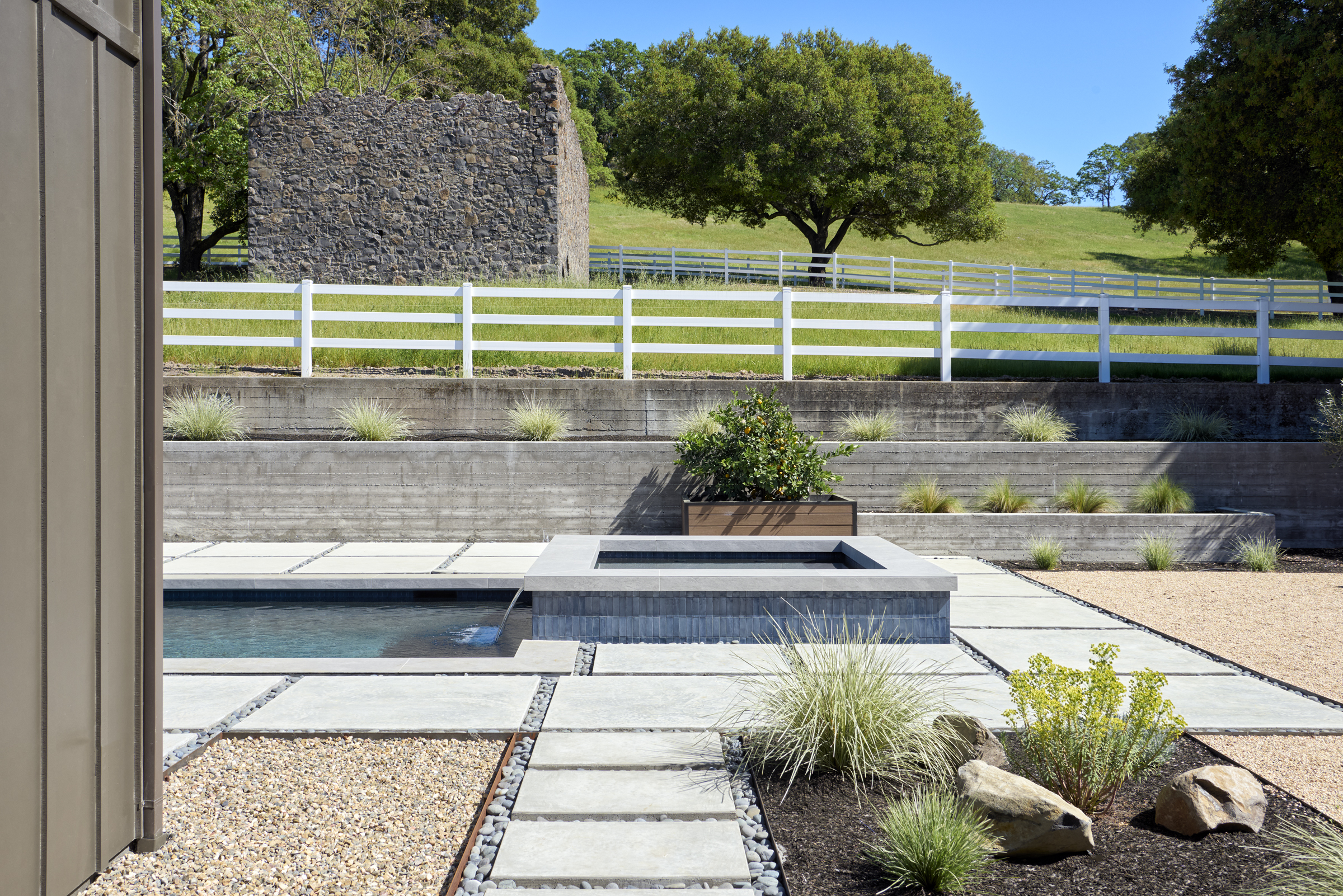 This modern outdoor living space features a sleek, rectangular swimming pool and an integrated spa with dark tile accents. The area is surrounded by clean concrete pavers, decorative gravel, and tiered retaining walls that lead up to a white fence and a historic stone ruin in the background. The composition offers a sophisticated, minimalist aesthetic that blends contemporary design with rustic landscape elements.