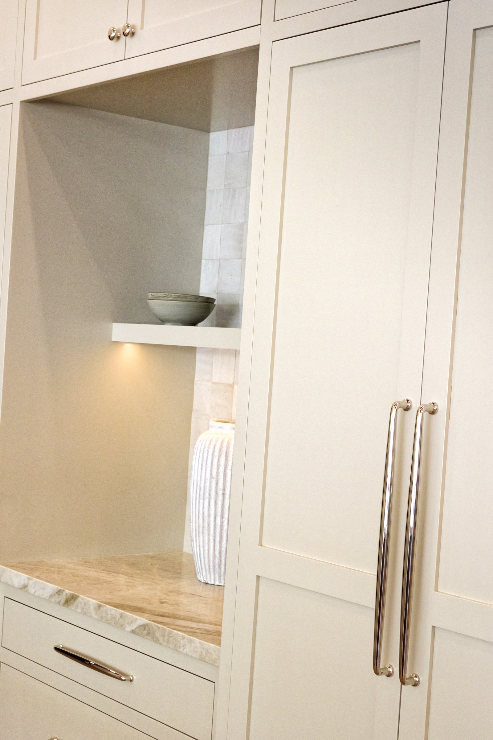 This image showcases a sophisticated, custom-built kitchen cabinetry unit featuring a recessed display niche with integrated under-shelf lighting. The design highlights high-end finishes, including a marble countertop, polished nickel hardware, and a textured tile backsplash, creating a luxurious and refined aesthetic. The perspective is a close-up, angled shot that emphasizes the craftsmanship and elegant material palette of the cabinetry.