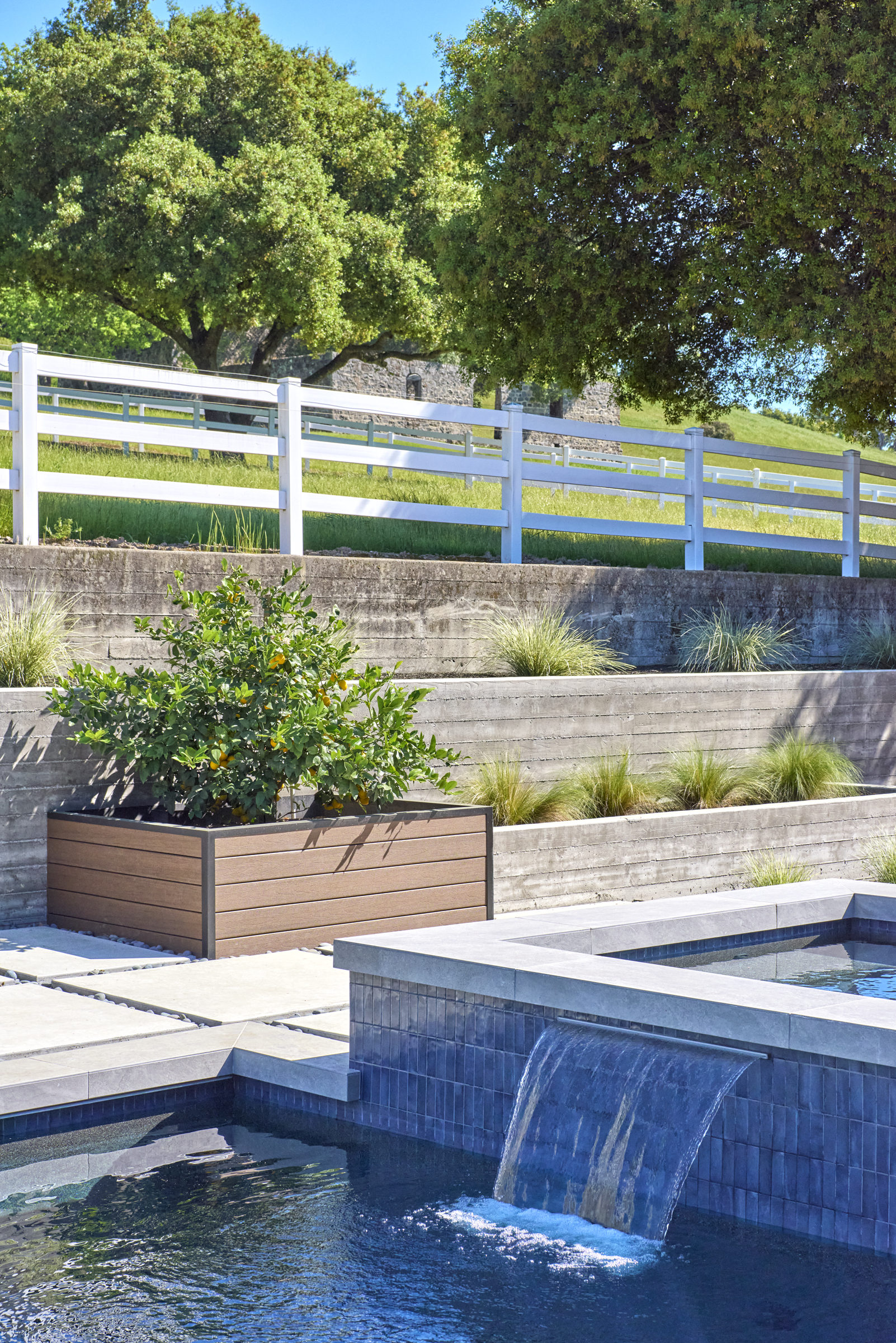 This serene outdoor space features a modern, dark-tiled spa with a cascading waterfall feature flowing into a pool. A sleek, wood-look planter box containing a citrus tree sits on a stone patio, set against a backdrop of tiered concrete retaining walls and a white ranch-style fence. The scene is bathed in bright, natural sunlight, creating a tranquil and luxurious backyard atmosphere.