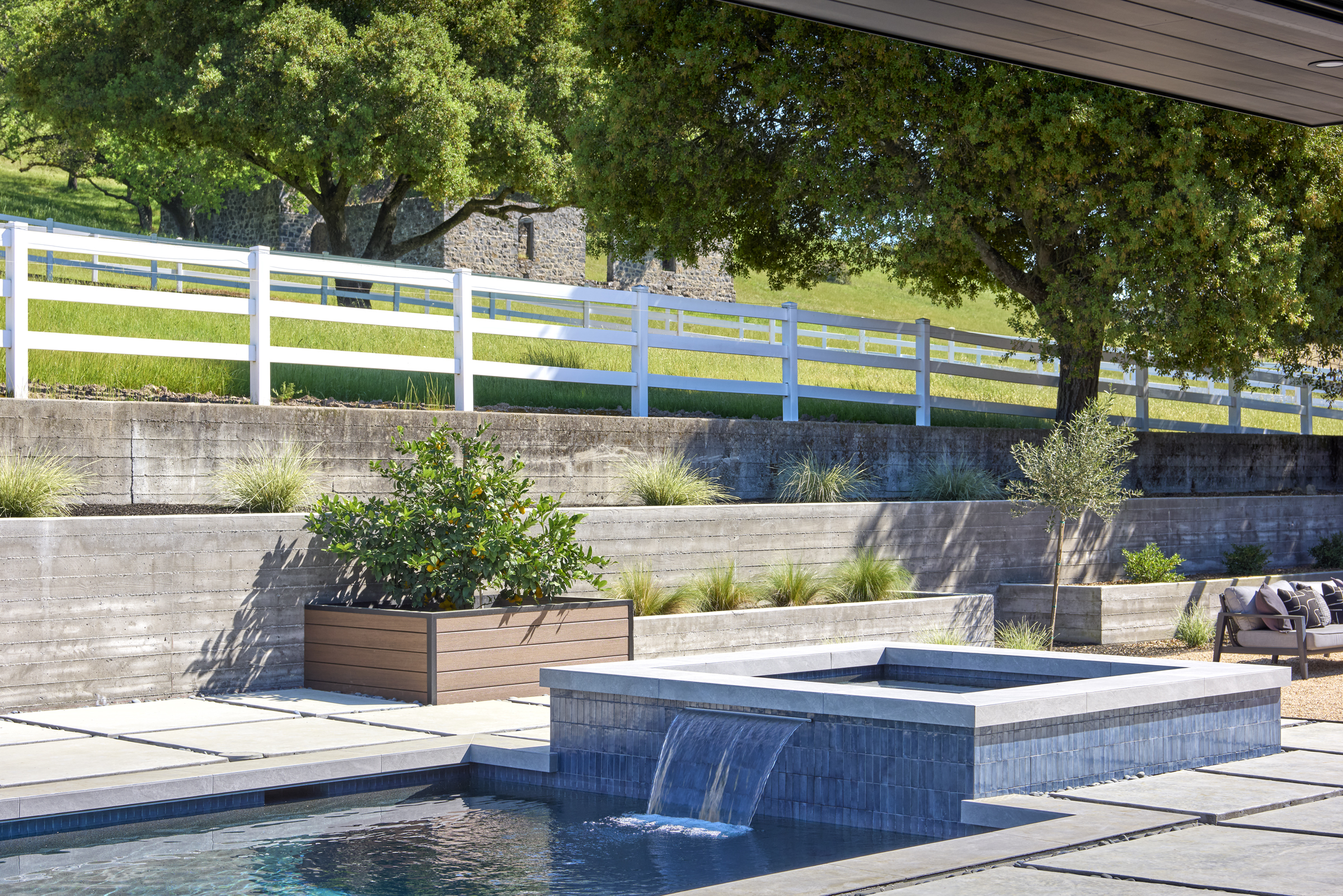 This elegant outdoor space features a modern, tiled spa with a cascading waterfall flowing into a swimming pool, set against a backdrop of tiered concrete retaining walls and lush landscaping. A large planter box with a citrus tree and a comfortable lounge area add to the sophisticated, resort-like atmosphere. The scene is framed by a white fence and mature trees, creating a serene and private backyard retreat.