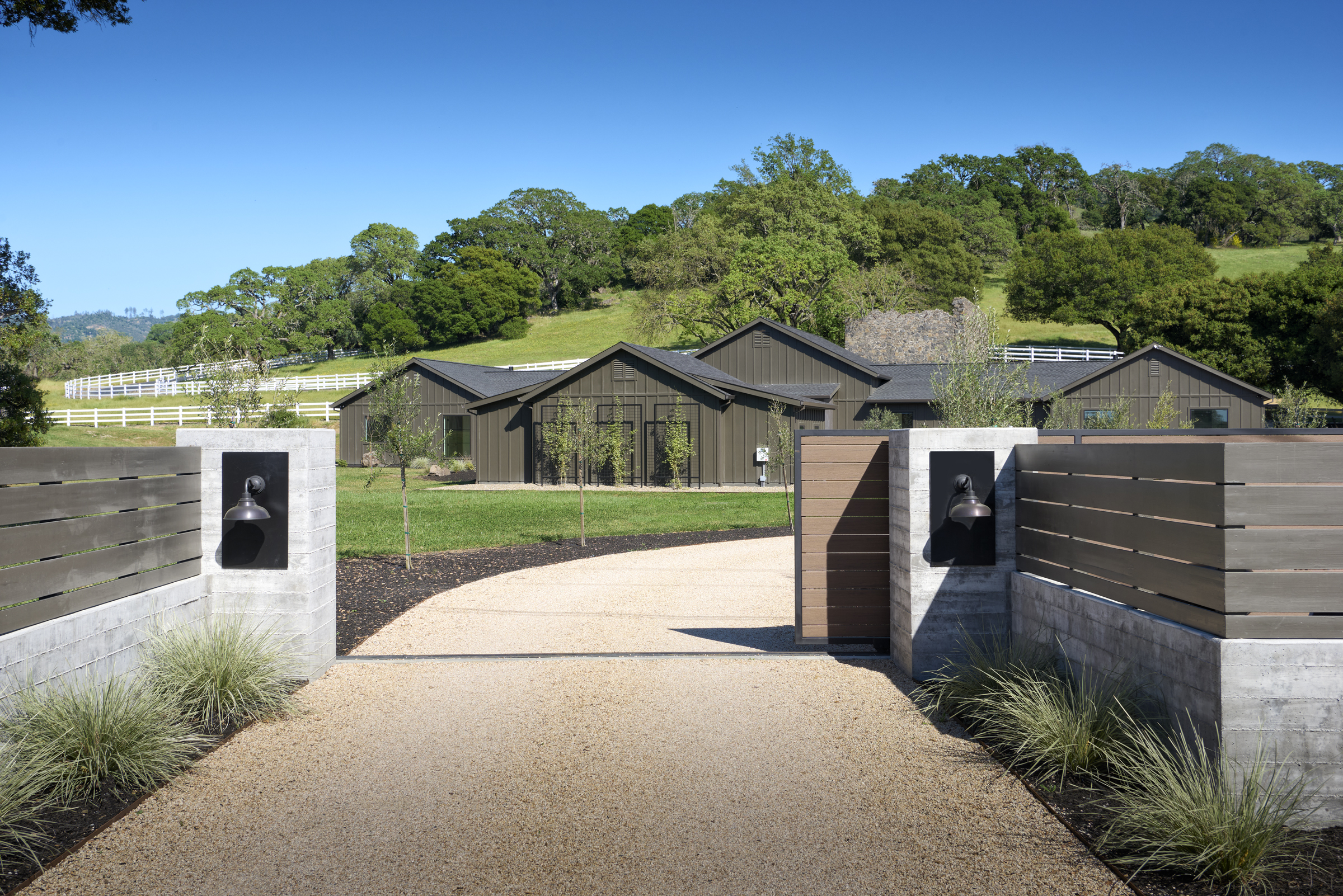 This perspective captures the grand entrance of a modern farmhouse-style estate, framed by concrete pillars and horizontal wood-slat gates. A gravel driveway leads the eye toward the dark-toned, multi-gabled residence set against a backdrop of lush green hills and mature oak trees. The scene conveys a sense of privacy, luxury, and seamless integration with the surrounding rural landscape.