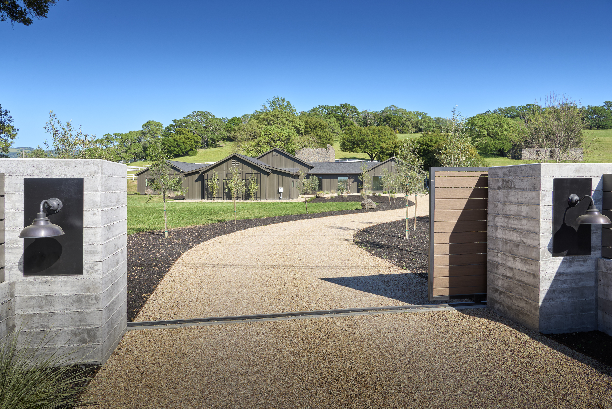 This perspective captures the grand entrance of a modern farmhouse estate, framed by two textured concrete pillars with sleek, dark metal sconces. A gravel driveway curves gracefully toward the dark-toned, multi-gabled residence, which is set against a backdrop of rolling green hills and mature trees. The scene conveys a sense of privacy, luxury, and architectural harmony between the contemporary structure and the natural landscape.