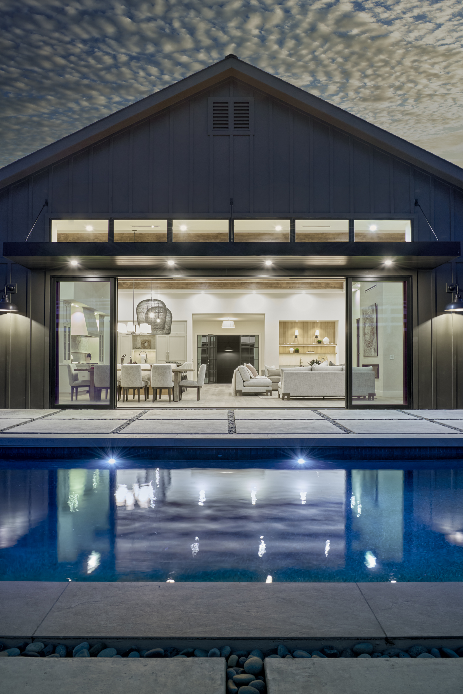 This stunning rear view of a modern farmhouse-style home showcases a seamless indoor-outdoor transition through expansive sliding glass doors that open to a serene swimming pool. The dark vertical siding contrasts beautifully with the warm, inviting interior lighting, highlighting a sophisticated open-concept living and dining area. The composition is perfectly symmetrical, emphasizing the clean lines of the architecture and the tranquil, high-end atmosphere of the backyard oasis.