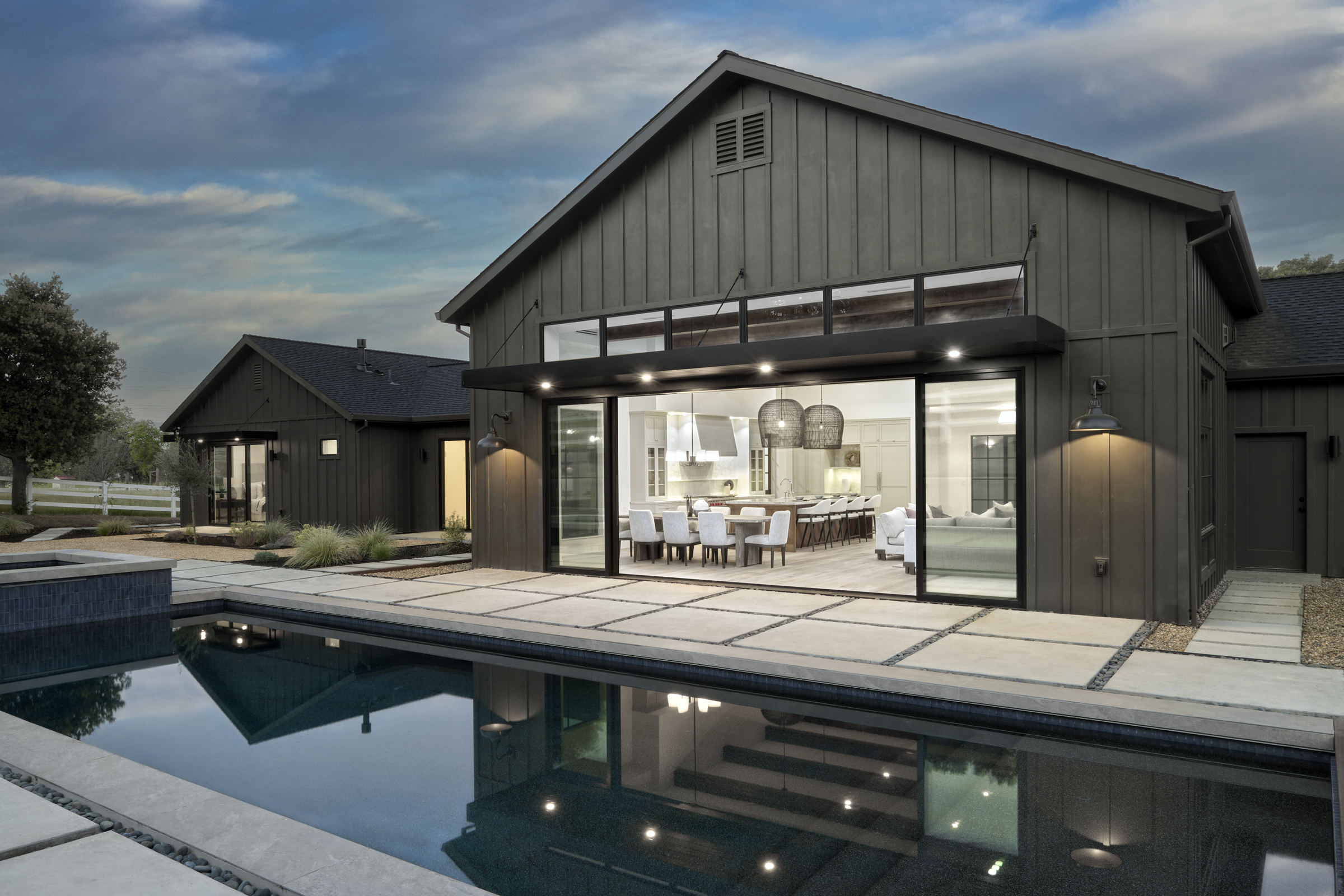 This stunning modern farmhouse features a seamless indoor-outdoor transition with expansive sliding glass doors opening from a bright, open-concept living and dining area onto a sleek concrete patio. The foreground is dominated by a dark-tiled, rectangular swimming pool that perfectly reflects the home's architectural lines and warm interior lighting. The dark vertical siding and clean, minimalist landscaping create a sophisticated, high-end aesthetic perfect for luxury outdoor living.