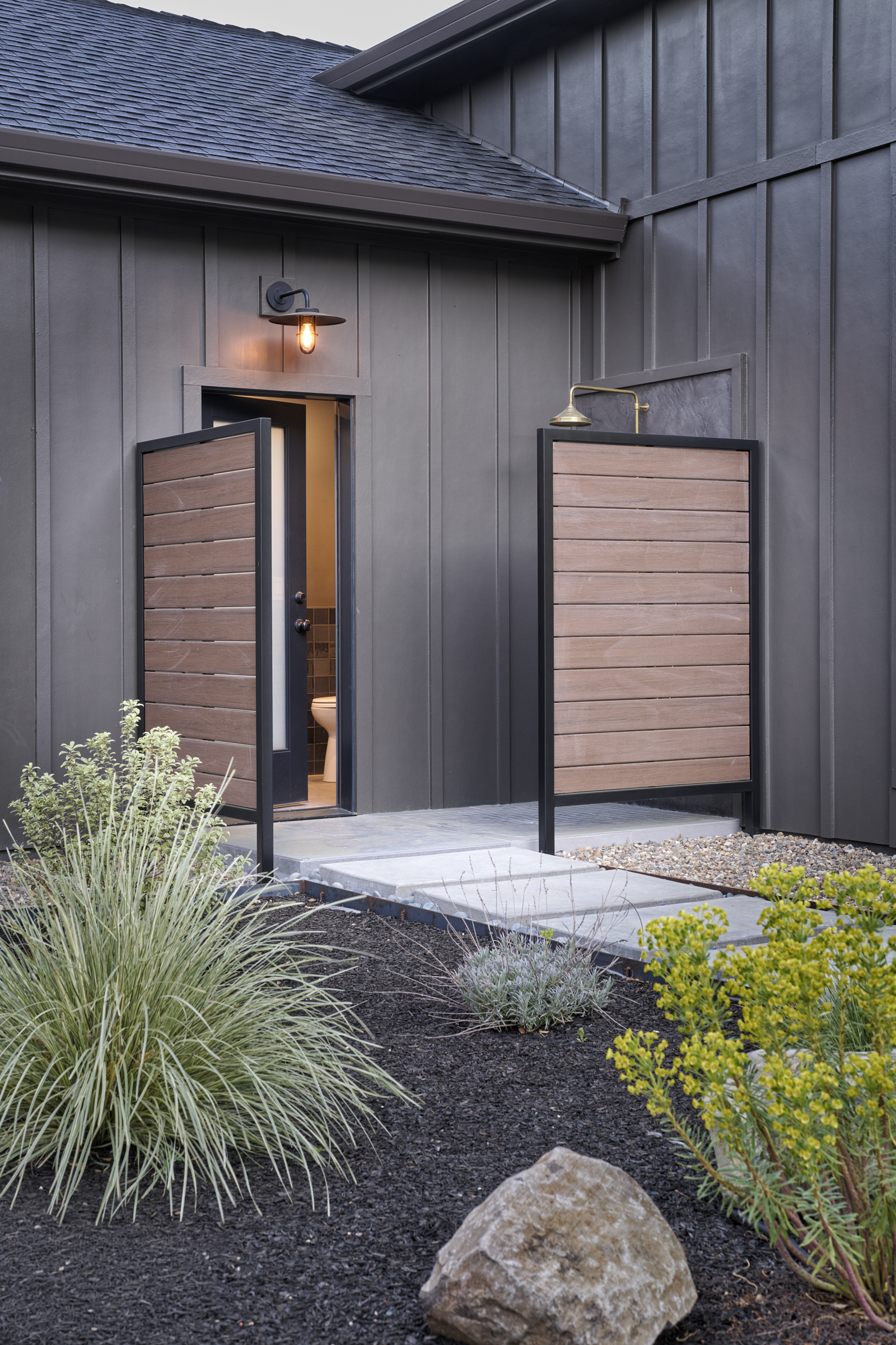 This modern exterior entryway features a striking dark vertical siding contrasted by warm wood-slat privacy screens and a minimalist outdoor shower setup. A concrete paver path leads to a doorway that reveals a glimpse of a bathroom interior, while a sleek wall-mounted light fixture adds a touch of industrial elegance. The surrounding landscaping, with dark mulch and ornamental grasses, enhances the contemporary, high-end aesthetic of the home.