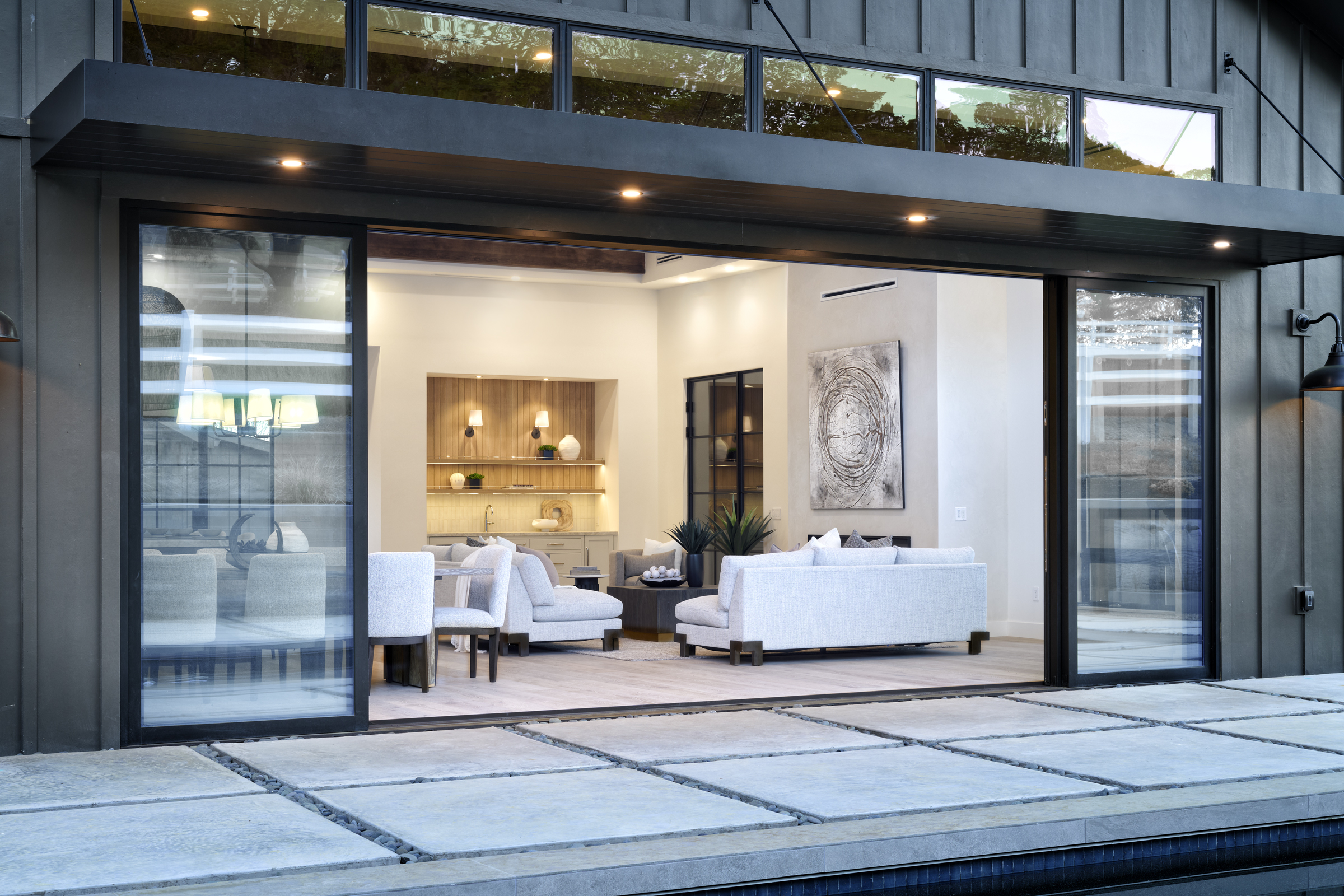 This image captures a seamless transition between a modern, dark-sided exterior and a bright, open-concept living space through expansive sliding glass doors. The foreground features a geometric concrete paver patio that leads directly into a sophisticated interior living area, complete with plush white seating and a built-in wet bar. The architectural design emphasizes indoor-outdoor living, creating a luxurious and inviting atmosphere perfect for entertaining.