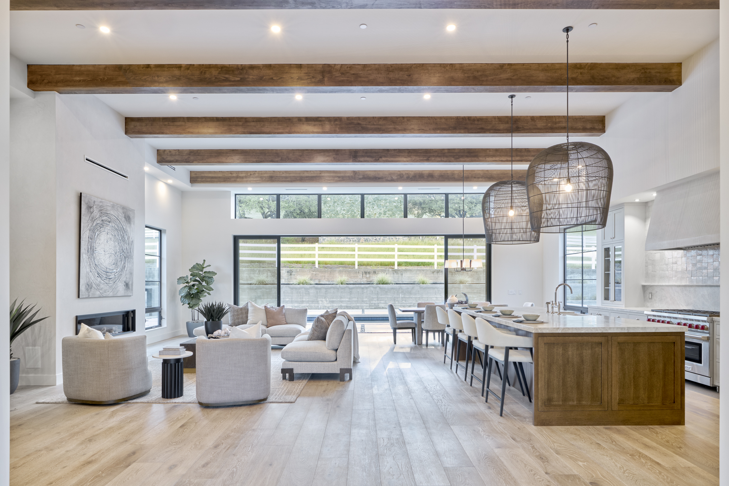 This expansive, open-concept living space seamlessly blends a modern living room with a gourmet kitchen, characterized by high ceilings with exposed wooden beams and light hardwood flooring. A large floor-to-ceiling glass wall provides a panoramic view of the outdoor landscape, while the interior features sophisticated neutral-toned furniture, oversized pendant lighting, and a sleek fireplace. The overall aesthetic is a refined, airy, and luxurious contemporary design perfect for high-end living.
