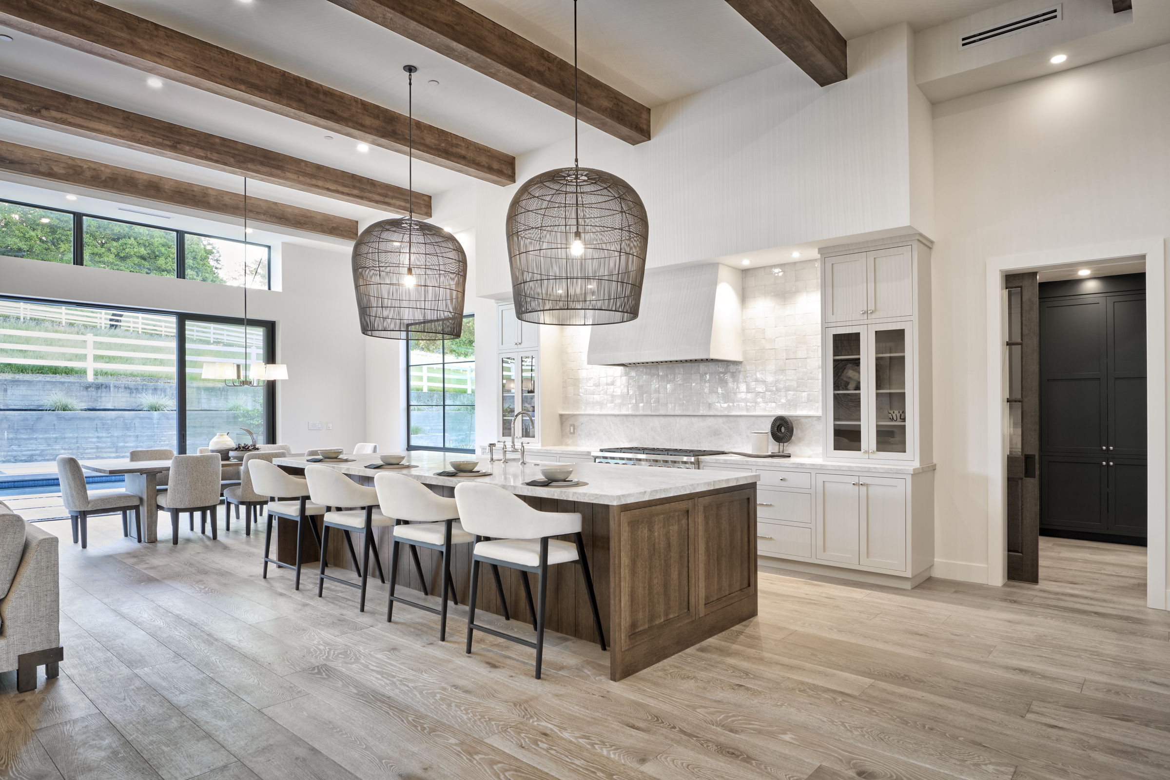 This expansive, modern kitchen features a large central island with a wood base and marble countertop, complemented by four sleek white bar stools. The space is defined by high vaulted ceilings with exposed wooden beams, elegant wire-mesh pendant lighting, and a seamless transition to the dining area and outdoor patio. The neutral color palette, high-end finishes, and open-concept layout create a bright, sophisticated, and inviting atmosphere perfect for entertaining.