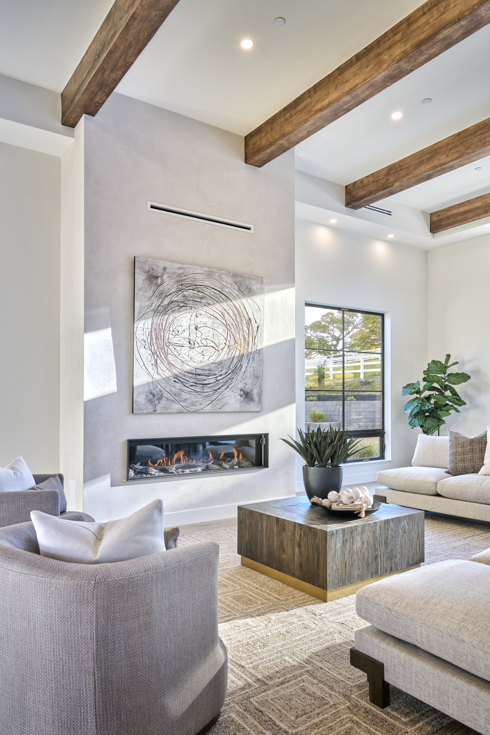 This sophisticated living room features a striking floor-to-ceiling fireplace with a modern linear insert and abstract artwork, framed by rustic wooden ceiling beams. The space is furnished with plush, neutral-toned seating and a substantial wooden coffee table, all set upon a textured area rug. Large windows provide a bright, airy atmosphere with a view of the outdoor landscape, creating a seamless blend of modern luxury and natural warmth.