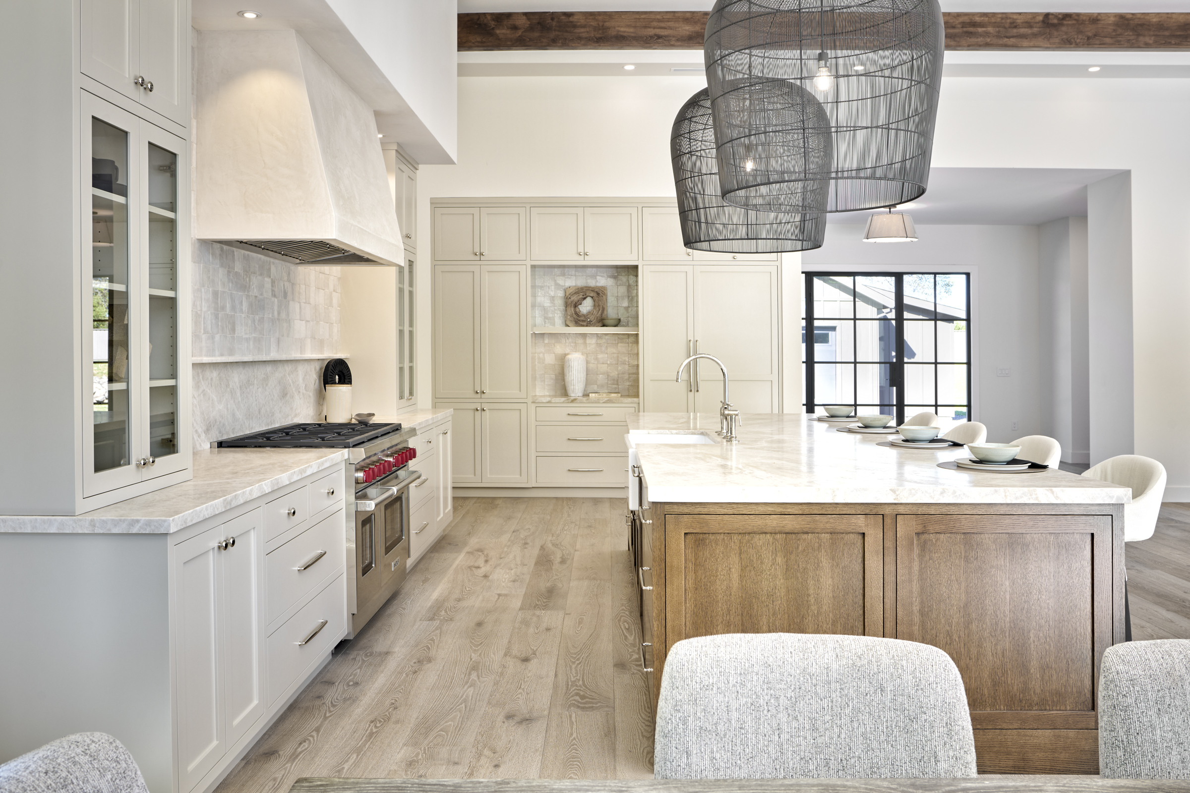 This sophisticated, high-end kitchen features a harmonious blend of light-toned cabinetry and a rich, wood-finished central island topped with a luxurious marble countertop. The space is anchored by a custom plaster range hood and dramatic, oversized wire-mesh pendant lights that draw the eye upward toward the exposed wooden ceiling beams. The open-concept design flows seamlessly into a bright dining area, creating an inviting and airy atmosphere perfect for both culinary tasks and social gatherings.