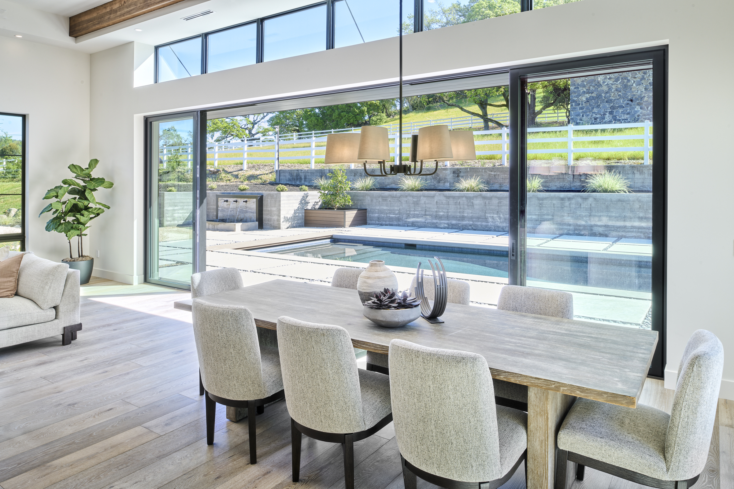 This elegant dining room features a large, rustic wooden table surrounded by upholstered chairs, creating a sophisticated yet inviting atmosphere. The space is defined by its seamless transition to the outdoors through expansive floor-to-ceiling sliding glass doors that reveal a pristine swimming pool and landscaped backyard. A modern chandelier hangs above the table, while the neutral color palette and clean lines emphasize a contemporary, high-end design aesthetic.