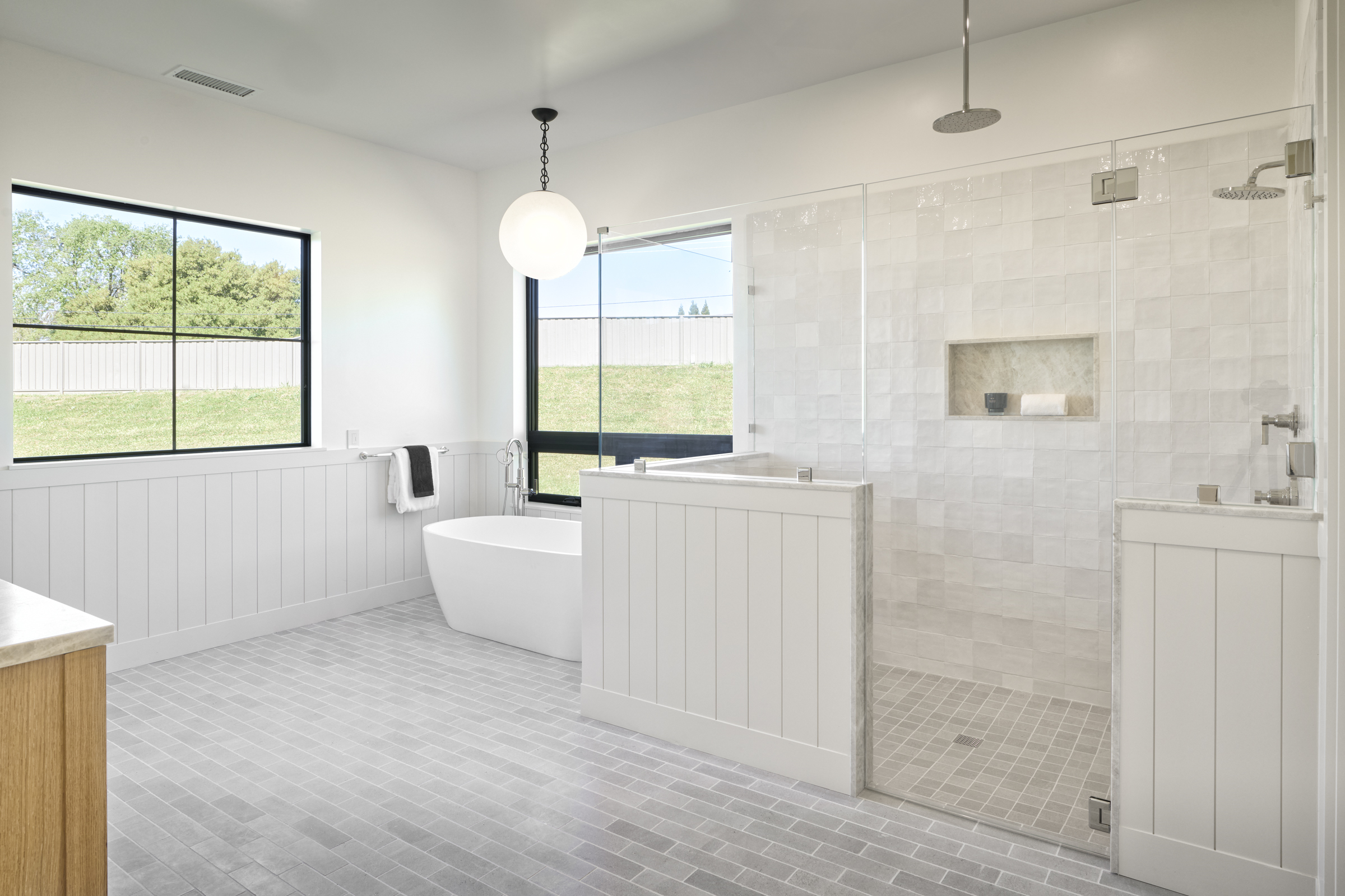 This bright and airy primary bathroom features a modern freestanding soaking tub positioned next to a large window, complemented by a spacious walk-in shower with white subway-style tiles and a glass enclosure. The room is finished with light gray floor tiles and white wainscoting, creating a clean, spa-like atmosphere. A minimalist globe pendant light hangs above the tub, adding a touch of contemporary elegance to the serene space.