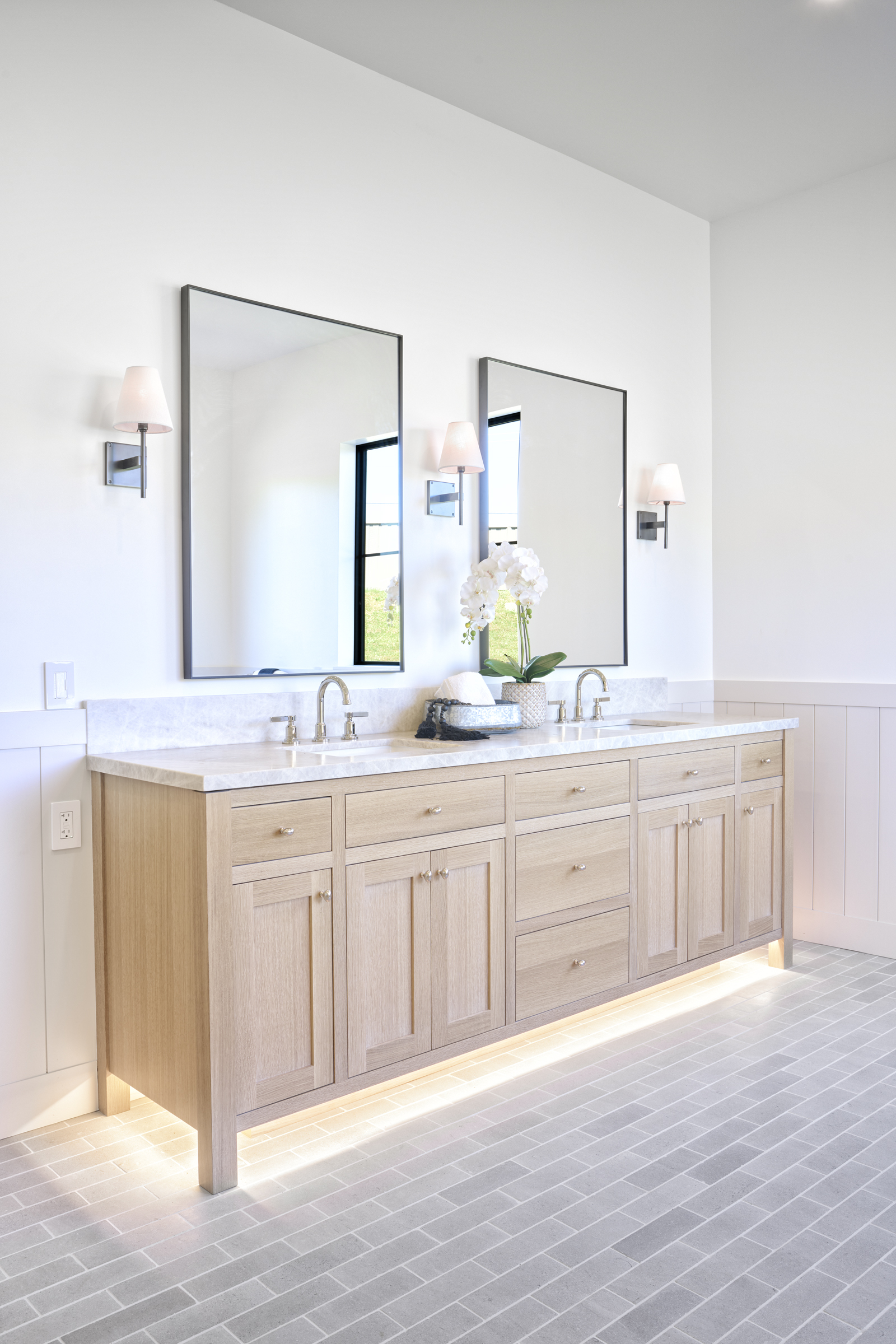 This elegant primary bathroom features a double vanity with light oak cabinetry, a marble countertop, and dual rectangular mirrors framed in dark metal. The space is illuminated by stylish wall sconces and a warm, under-cabinet LED glow that highlights the grey brick-style floor tiles. The clean, minimalist design and neutral color palette create a bright, sophisticated, and spa-like atmosphere.